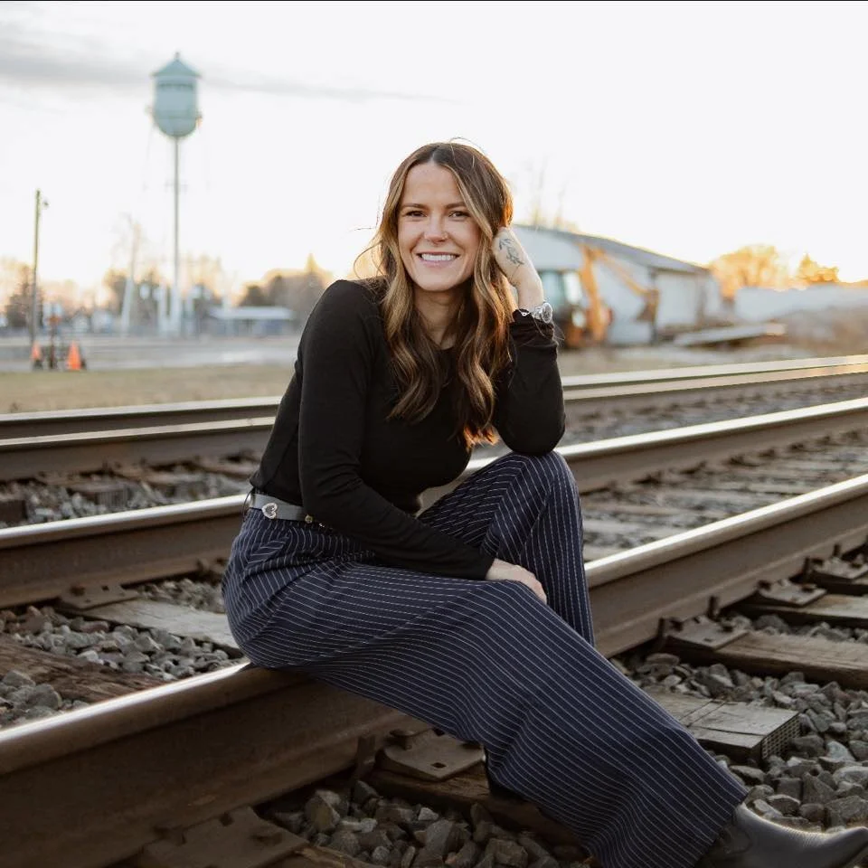 A woman with long hair, wearing a black top and pinstriped pants, sitting on train tracks during sunset, smiling at the camera.