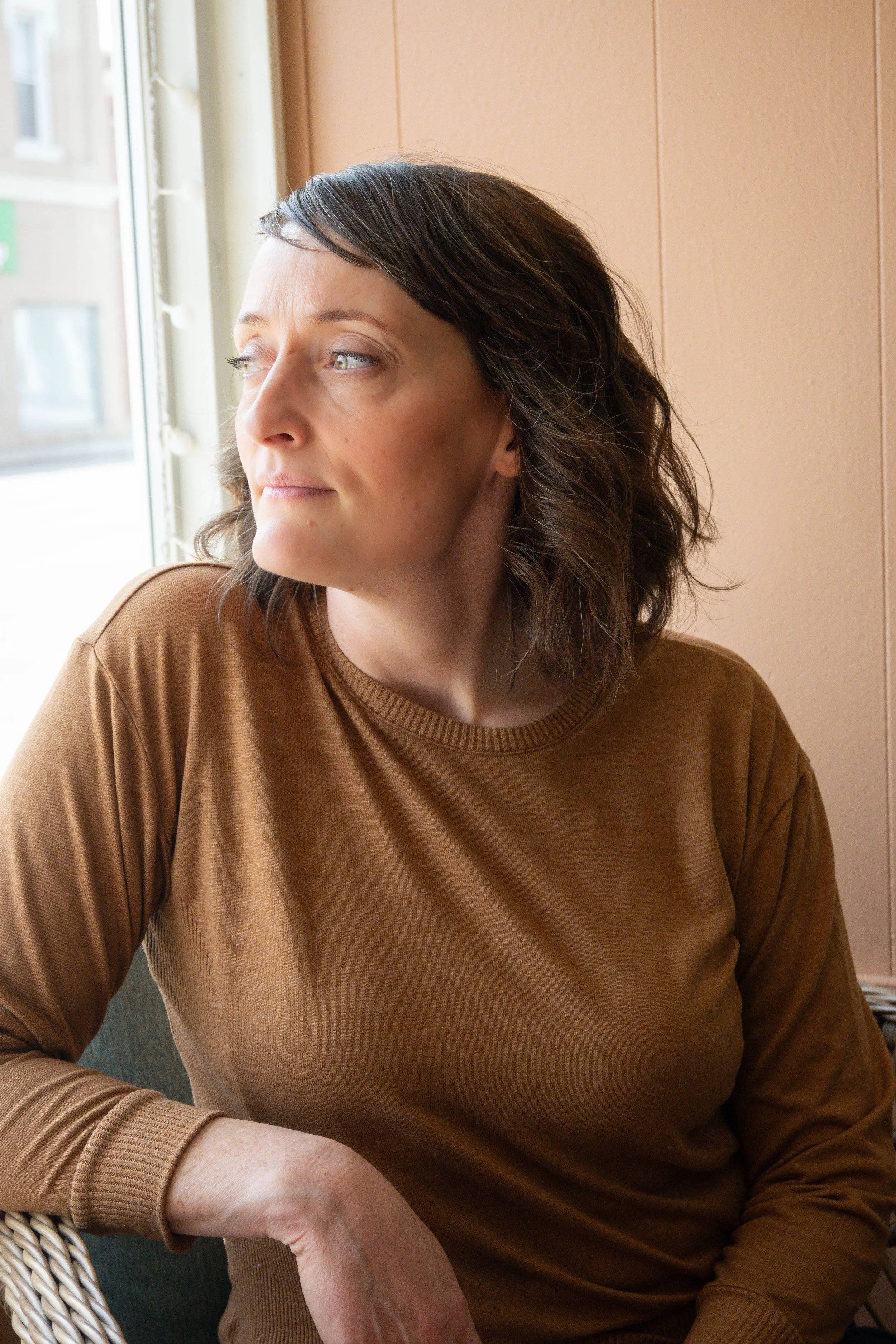 A woman with short dark brown hair looking out a window, wearing a brown long-sleeve shirt.