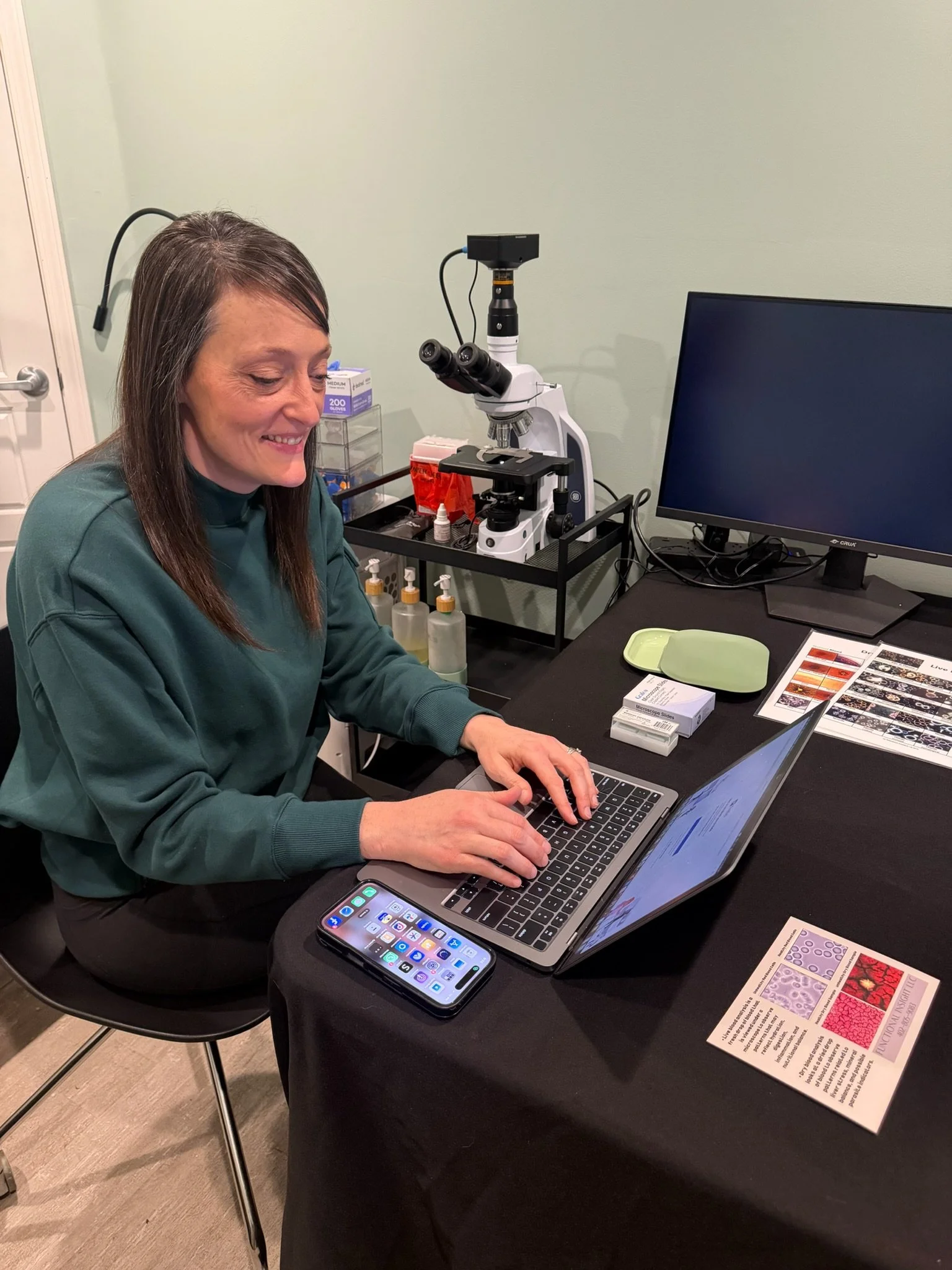 Woman using a laptop at a desk with a microscope and a smartphone nearby.