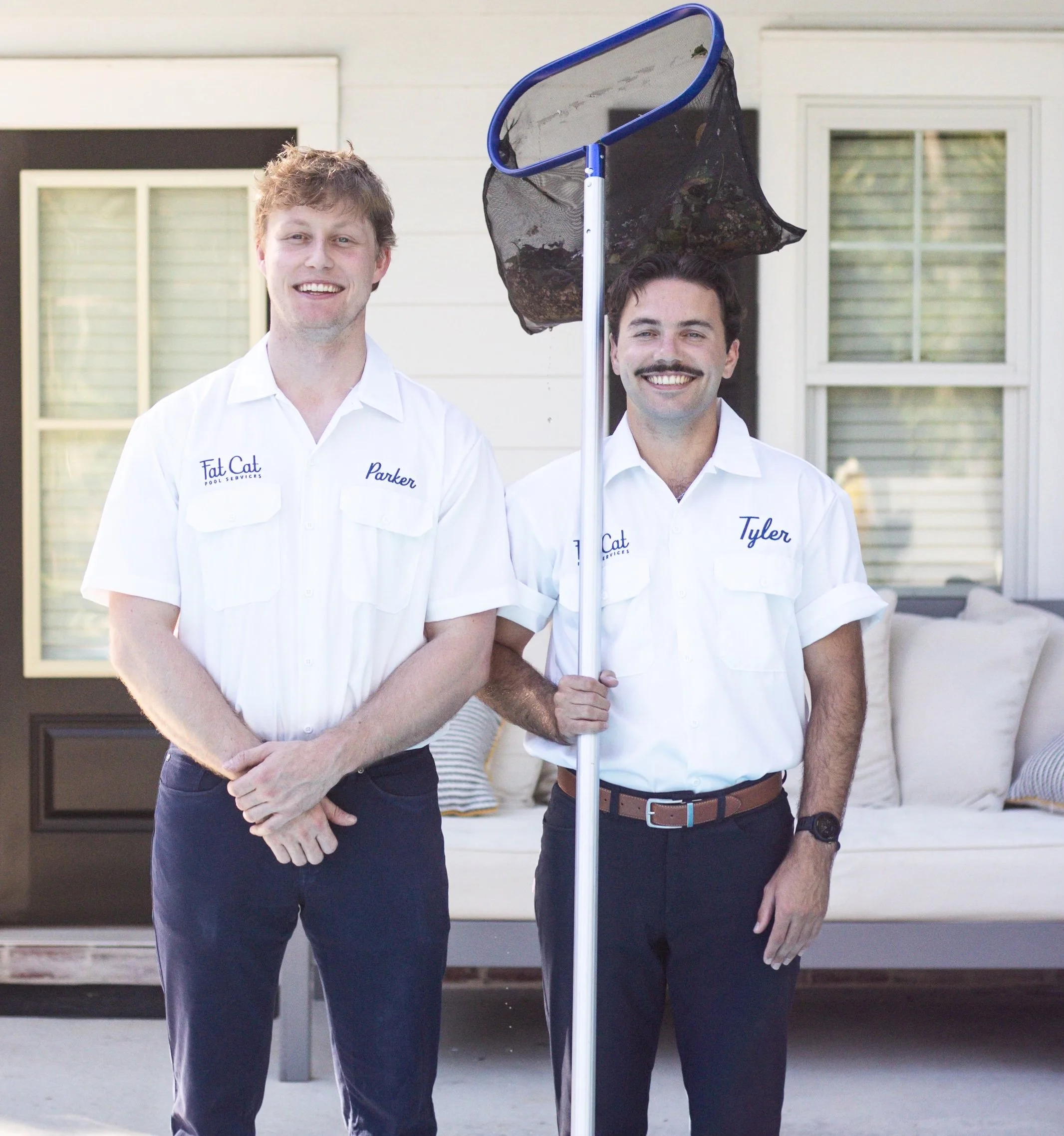 Two men standing outside in front of a house, holding a pool skimmer net with leaves and debris. They are wearing white shirts with embroidered names, smiling at the camera.