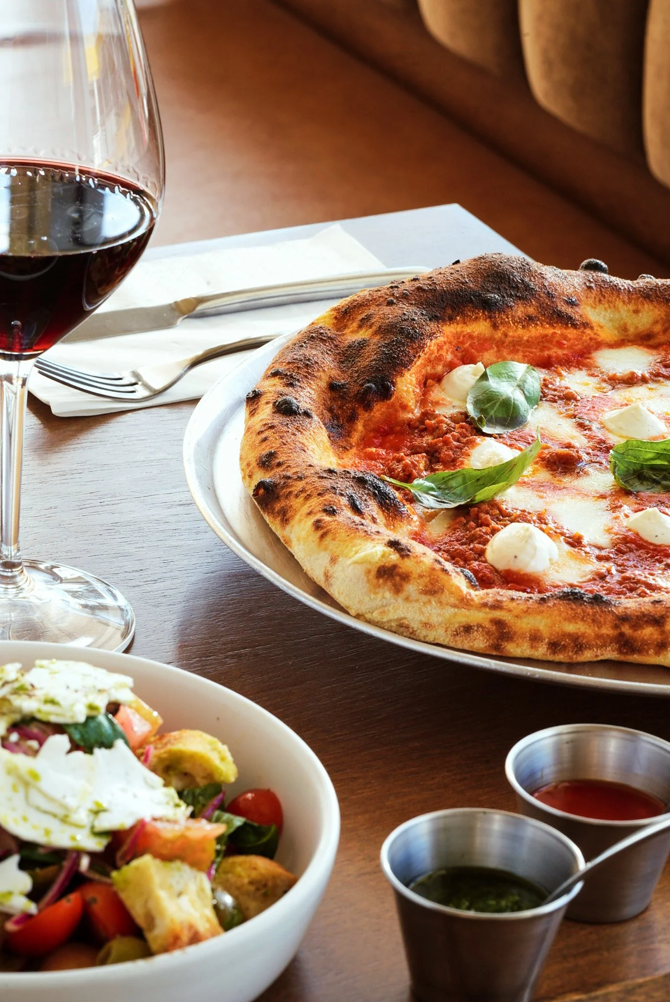 Una pizza de queso y albahaca, una copa de vino tinto, y una ensalada en un comedor de restaurante.