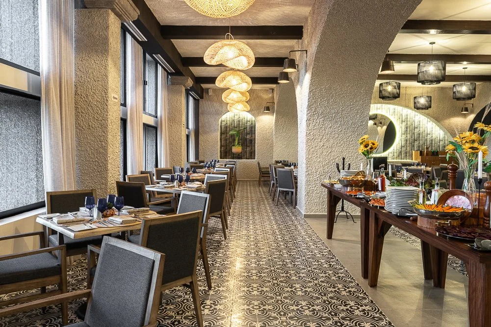 Sala de restaurante elegantemente decorada con mesas, sillas, ventanas grandes y luz ambiental suave. Hay una mesa de buffet con platos y flores decorativas.