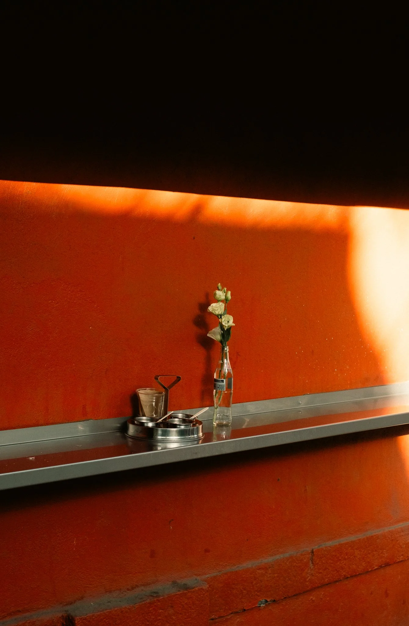 Vaso con flores blancas en una superficie metálica junto a utensilios de cocina, en una pared de color naranja.
