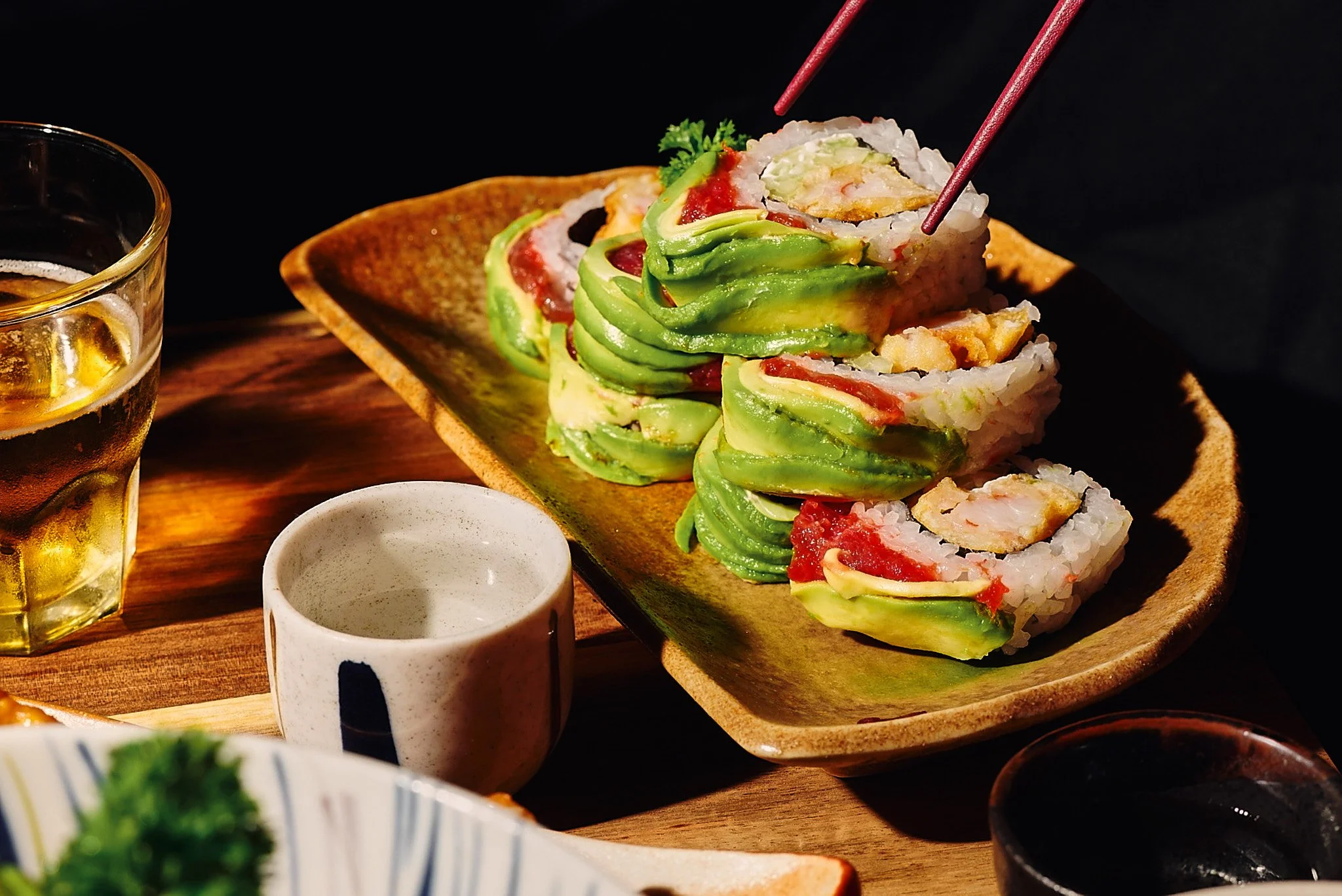 Rollos de sushi con aguacate y cangrejo, acompañados de salsa y bebida, en un plato de cerámica sobre una mesa de madera.