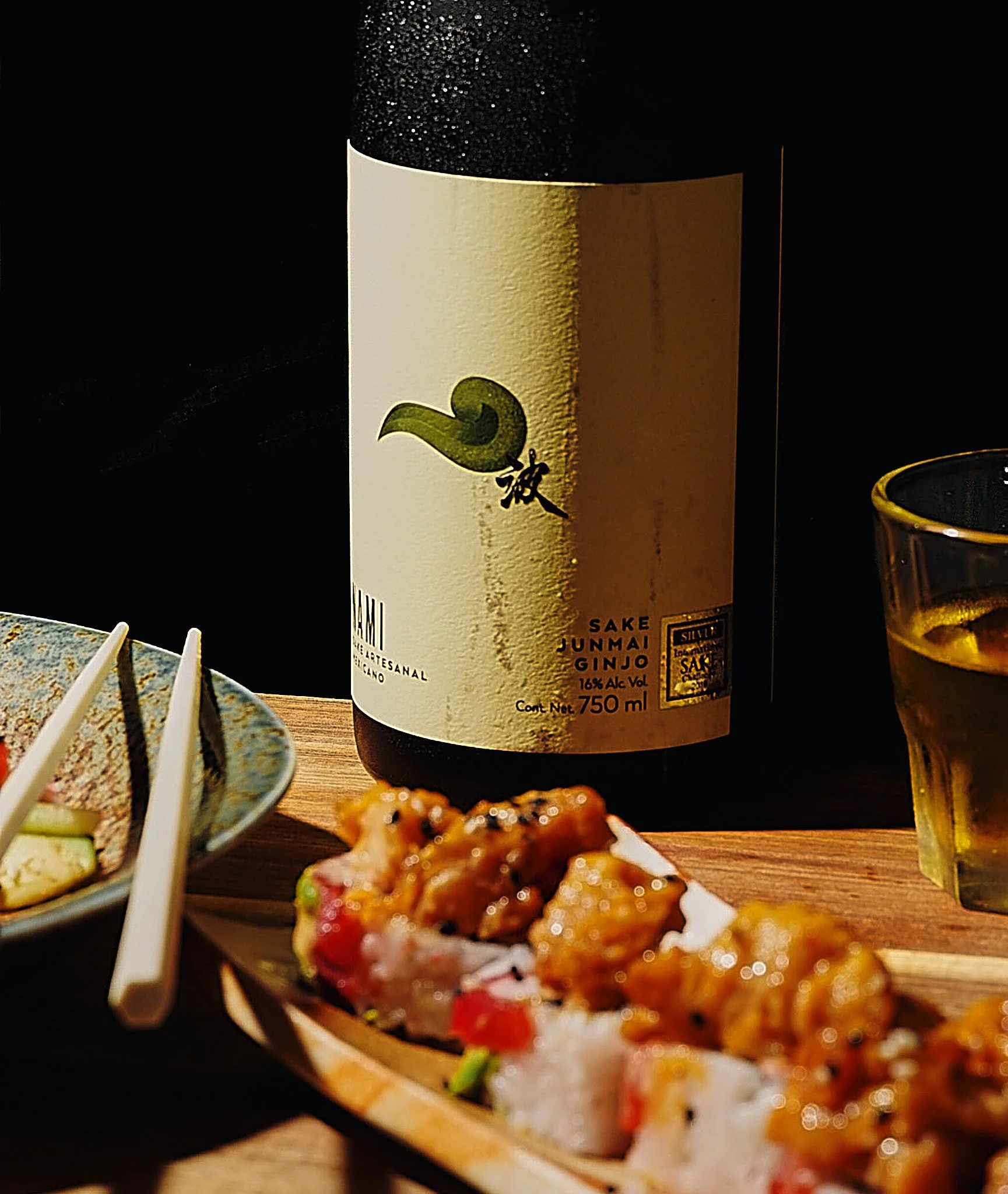 Botella de sake junmai ginjo de 750 ml, plato de comida japonesa con sushi y palillos a la izquierda, vaso de licor y una pieza de sushi con salsa en primer plano.