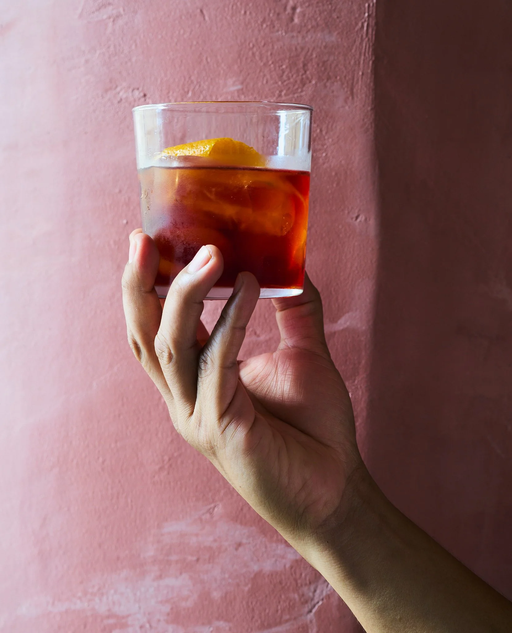 Mano sosteniendo un vaso con bebida refrescante, hielo y una rodaja de limón, contra una pared rosa.