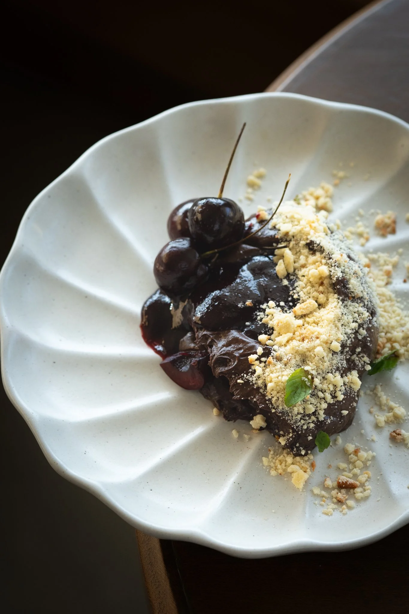 Postre cubierto de salsa de chocolate con cerezas y polvo de queso o galleta, en plato blanco con diseño decorativo.