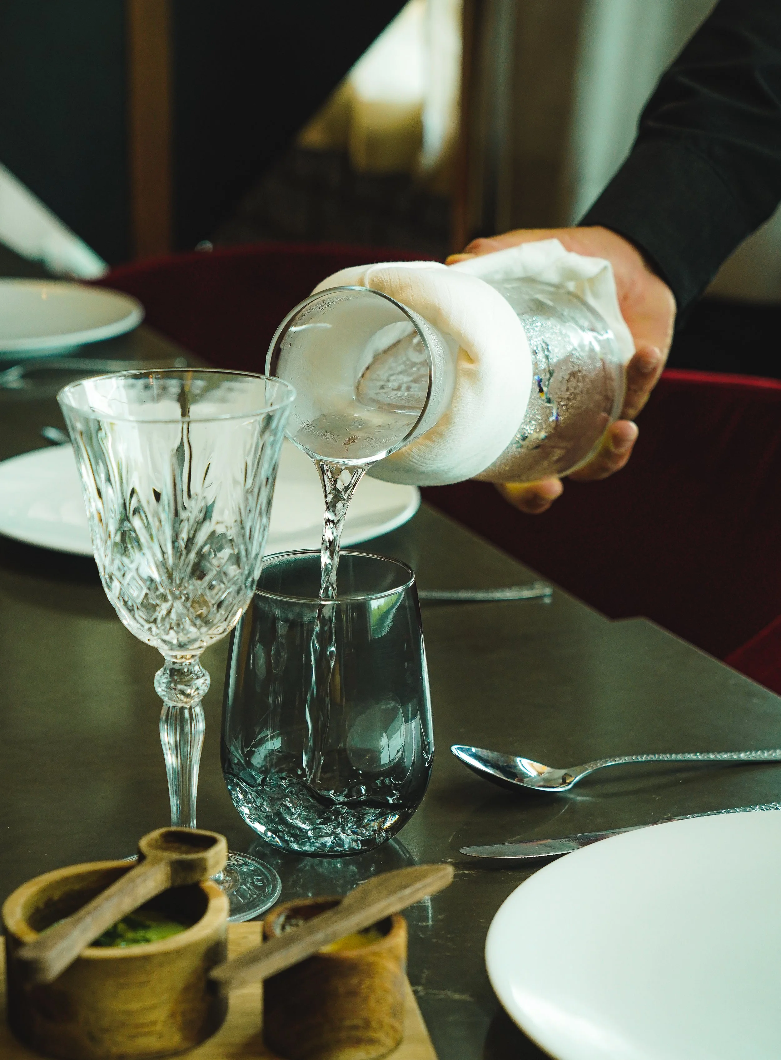 Una persona sirviendo agua en un vaso en un comedor elegante con cristalería y utensilios de plata.