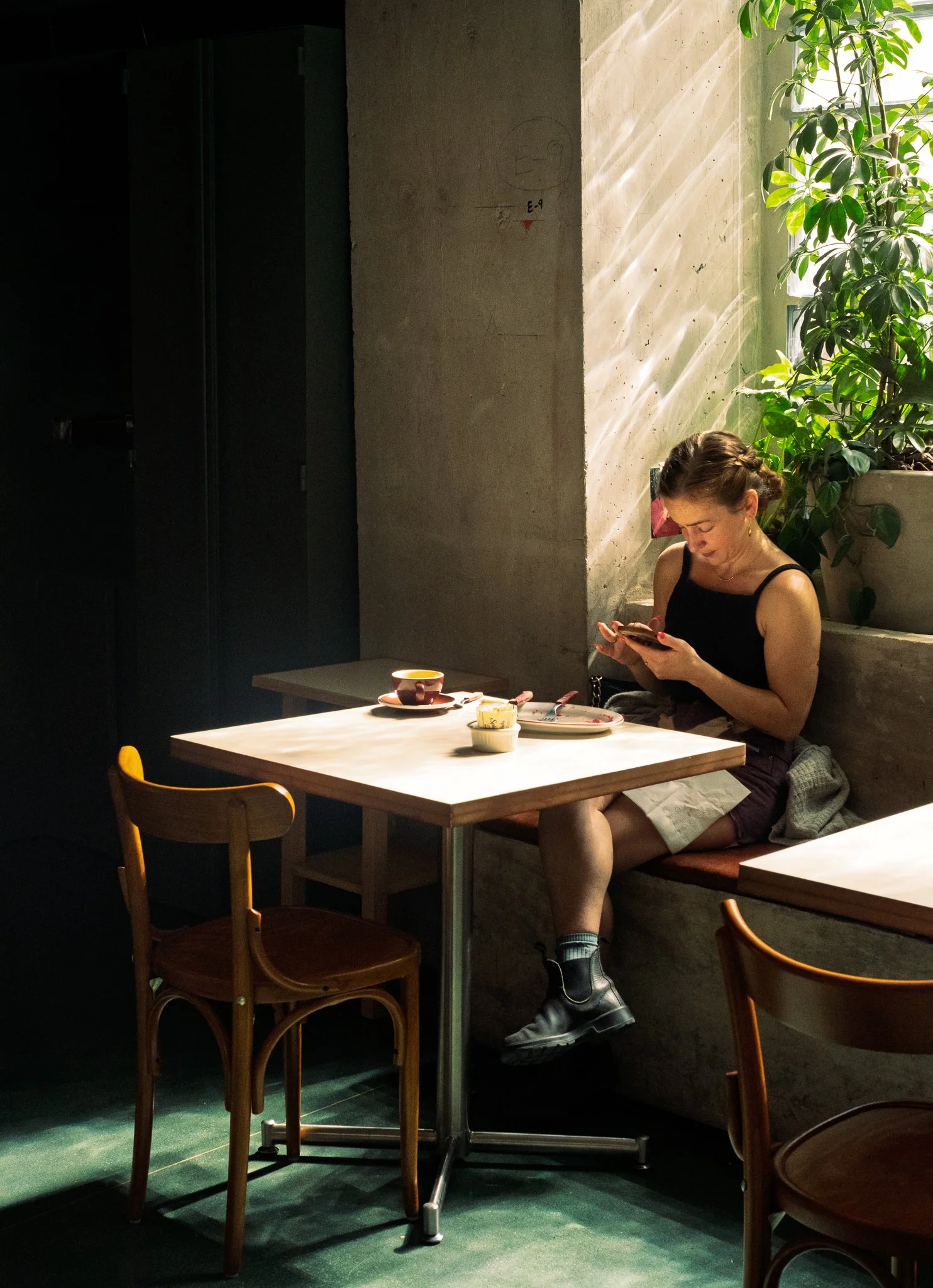 Mujer joven con rastas usando ropa negra, sentada en un restaurante o cafetería, mirando su teléfono móvil. Al fondo hay plantas verdes y una pared de cemento con rayos de luz natural entrando por una ventana.