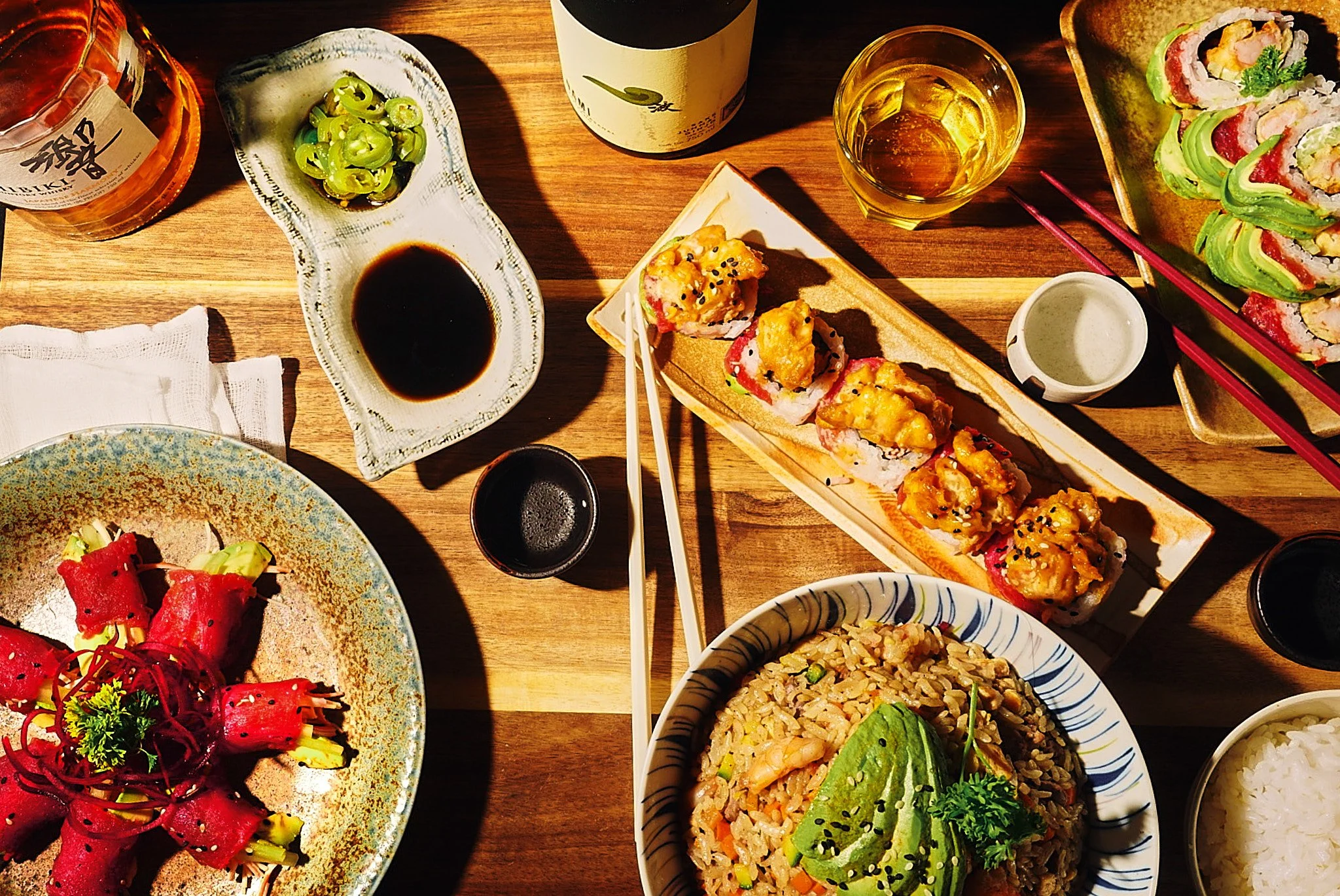 Imagen de una mesa con comida japonesa, incluyendo sushi, arroz frito, ensalada de remolacha, salsas, té y vino, en un ambiente con luz cálida.