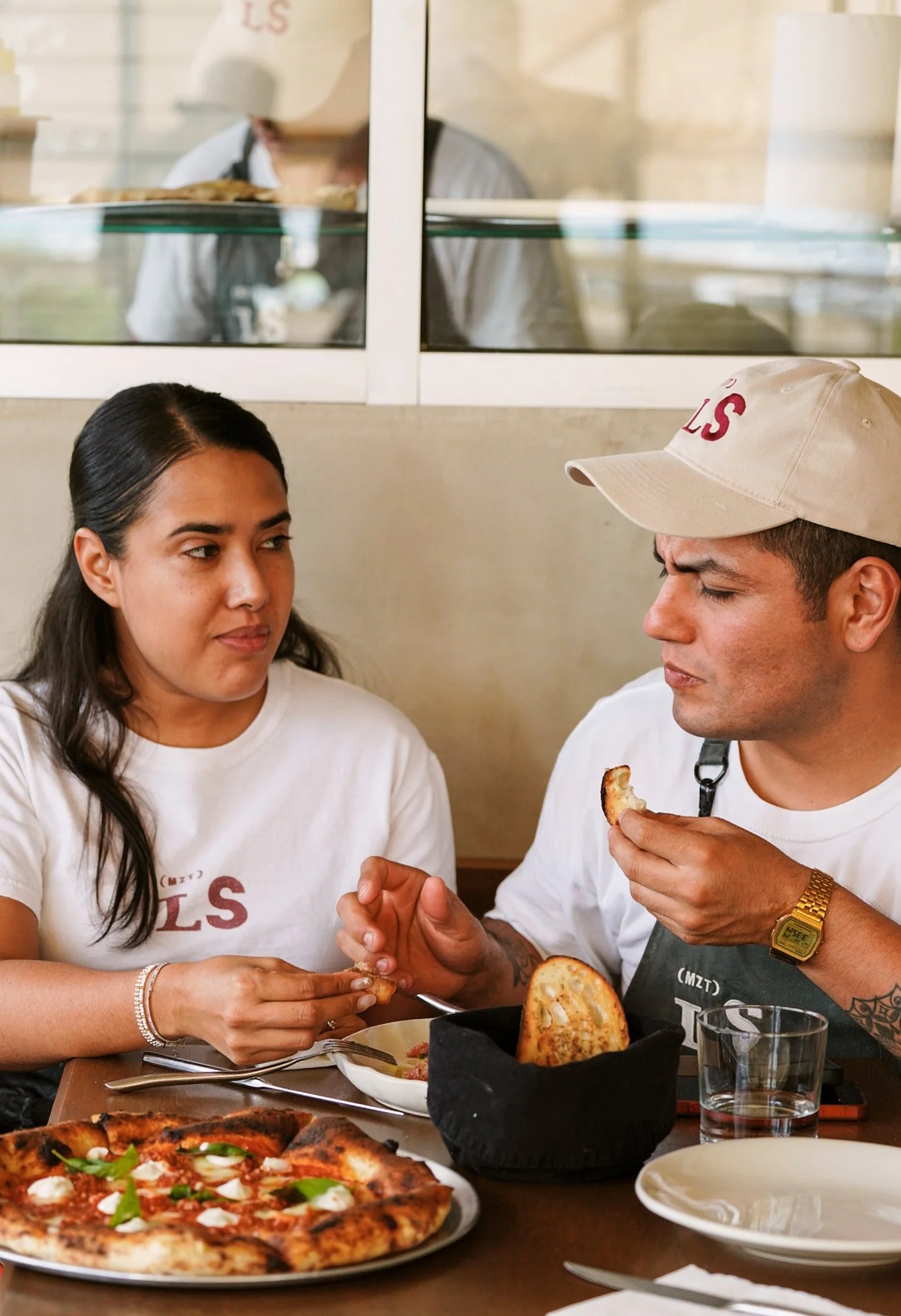 Dos personas sentadas en un restaurante compartiendo pan y pizza, con un camarero sirviendo en el fondo.