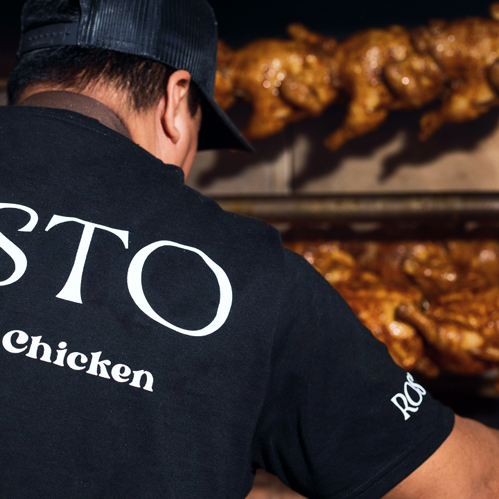 A man wearing a black cap and black shirt labeled 'ROSTO Chicken' is grilling or preparing rotisserie chicken.