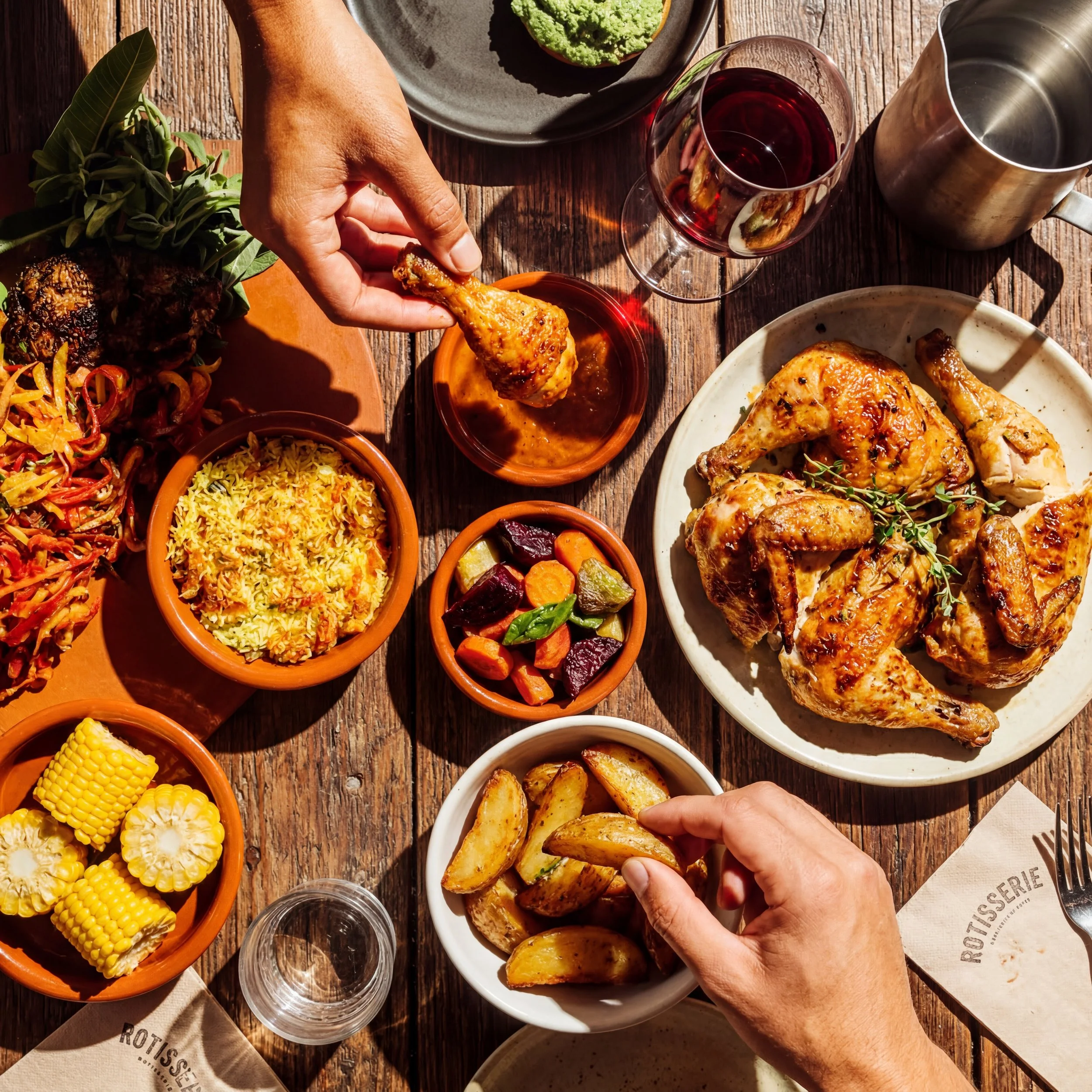 A family dinner filled with various dishes, including roasted chicken, roasted potatoes, mixed vegetables, rice, and salad. Two hands are reaching for chicken and potatoes. There is a glass of red wine and a metal pitcher.