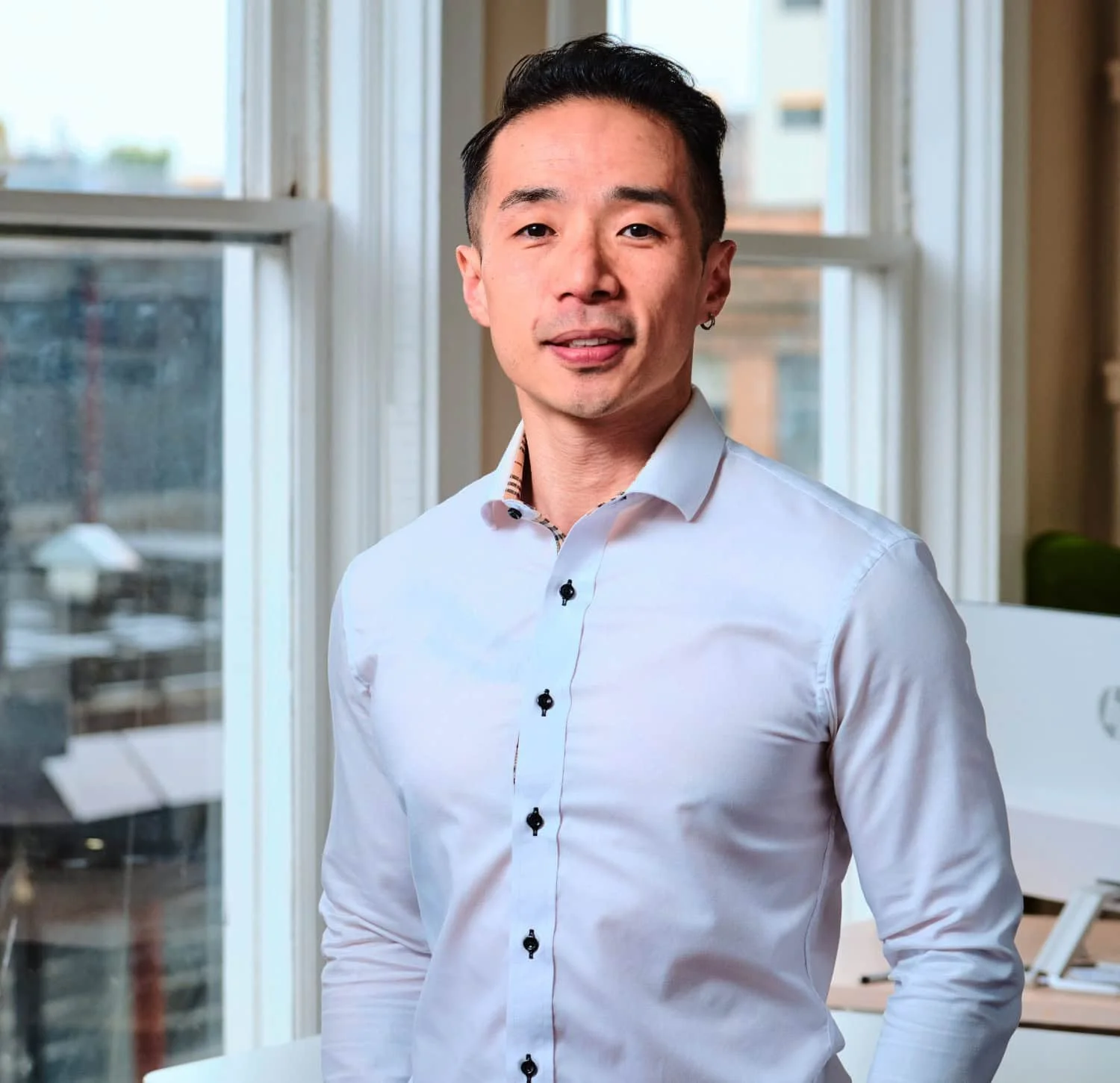 Vincent Lo. Building Performance Analyst Dip.T at reLoad Sustainable Design Inc. A man with short dark hair, wearing a white dress shirt with decorative buttons, standing in a bright office near large windows with Vanocuver views.