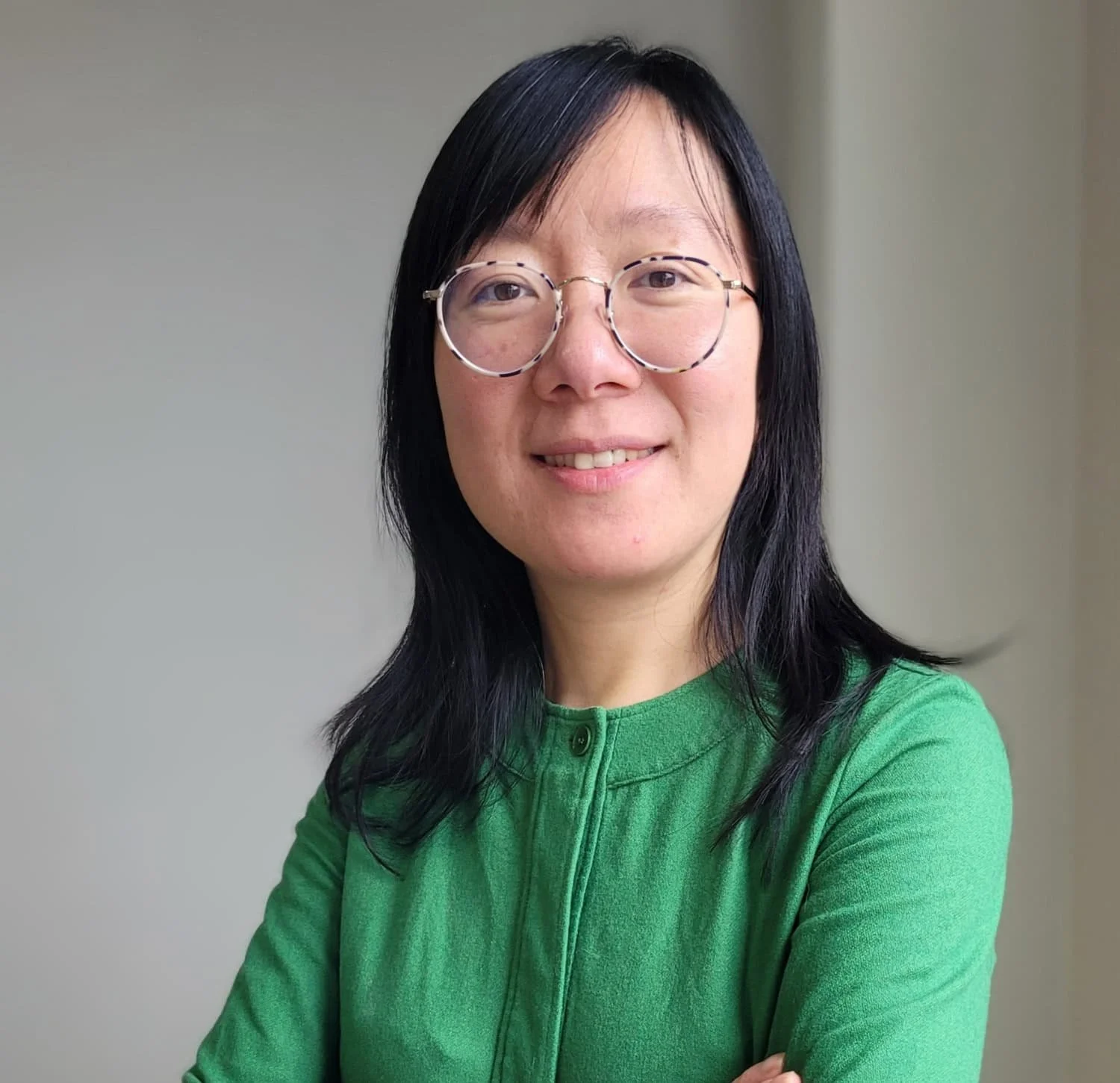 Ruffy Ruan. Project Manager + Building Performance Analyst  P.Eng., CPHC at reLoad Sustainable Design Inc.  Portrait of a woman with black hair and glasses, smiling, wearing a green top, standing indoors against a plain wall.