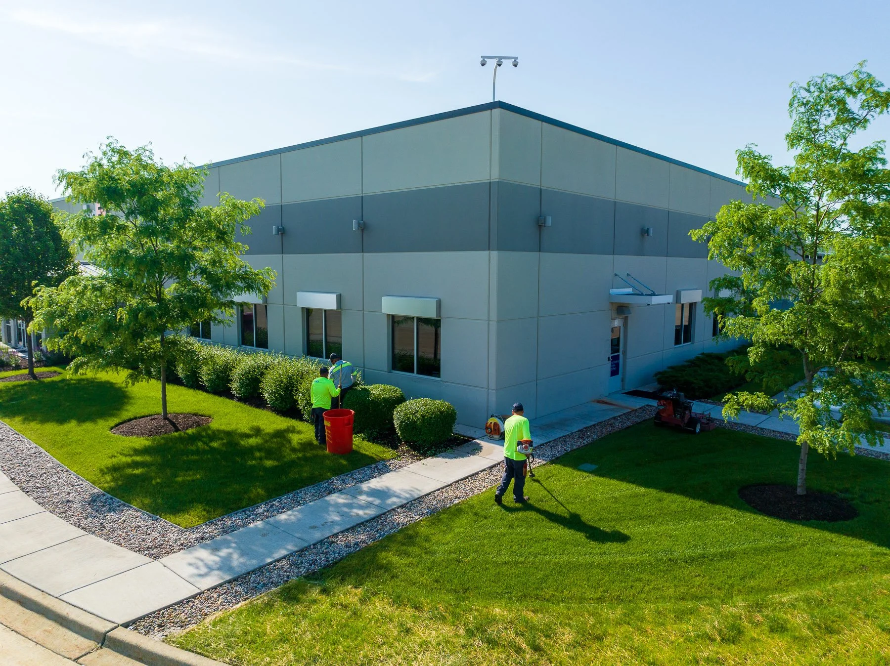 Workers trimming bushes outside a commercial building with a manicured lawn and trees.