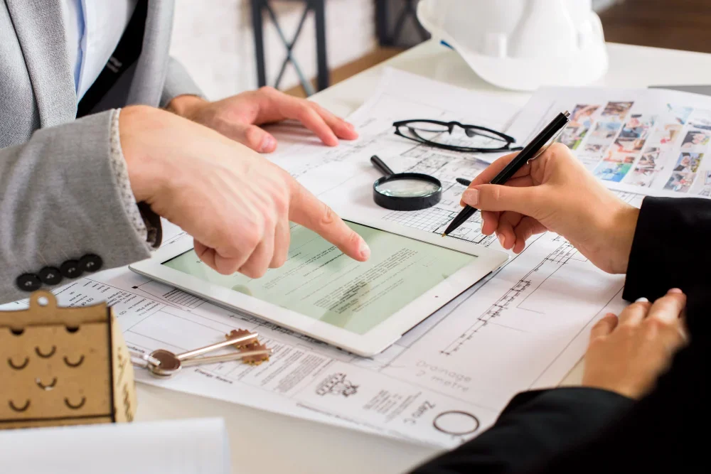 Two people review documents and a digital tablet on a desk with eyeglasses, a magnifying glass, photos, and stationery.