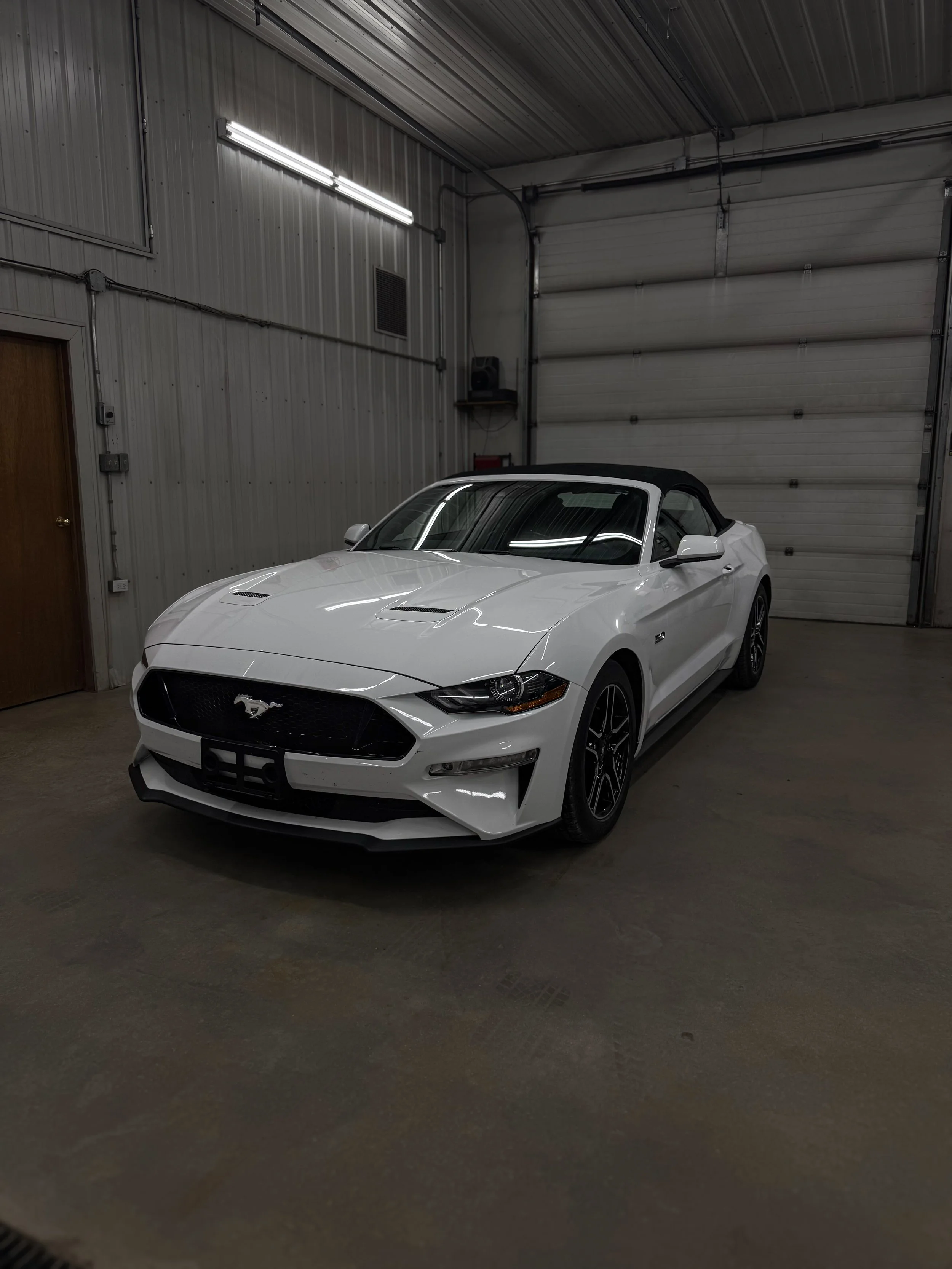 White Ford Mustang convertible parked inside a garage with metal walls and a large roll-up door.