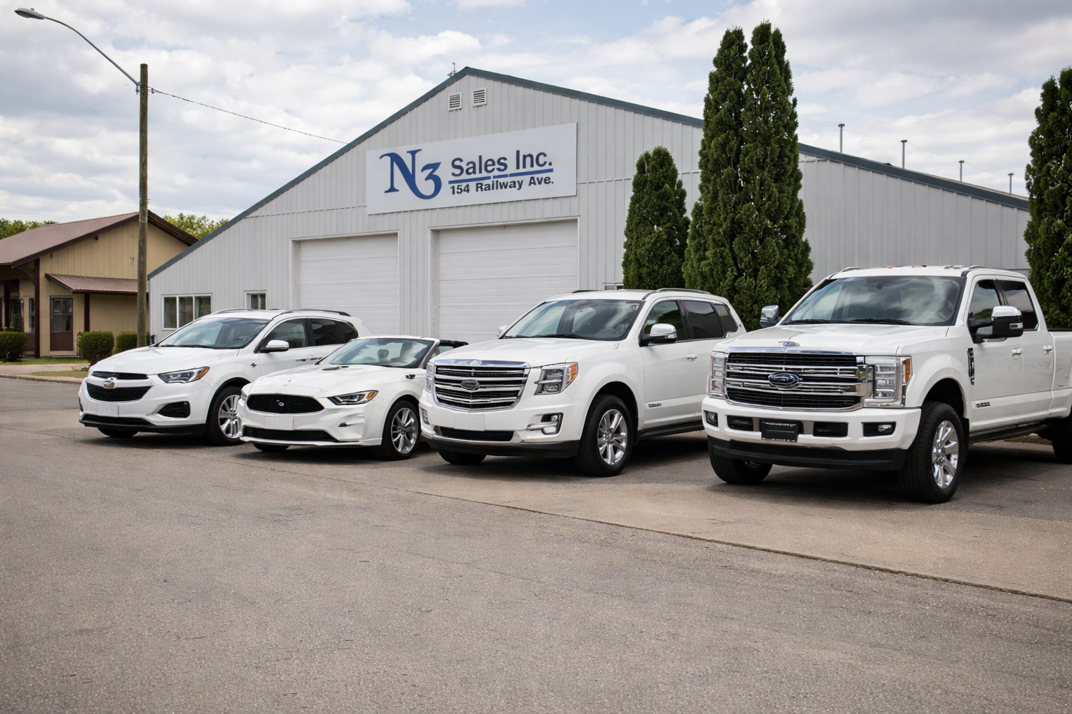 Four white vehicles parked in front of a white building with a sign that reads 'N3 Sales Inc., 154 Railway Ave.' Trees are visible behind the building, and a residential house is seen to the left.