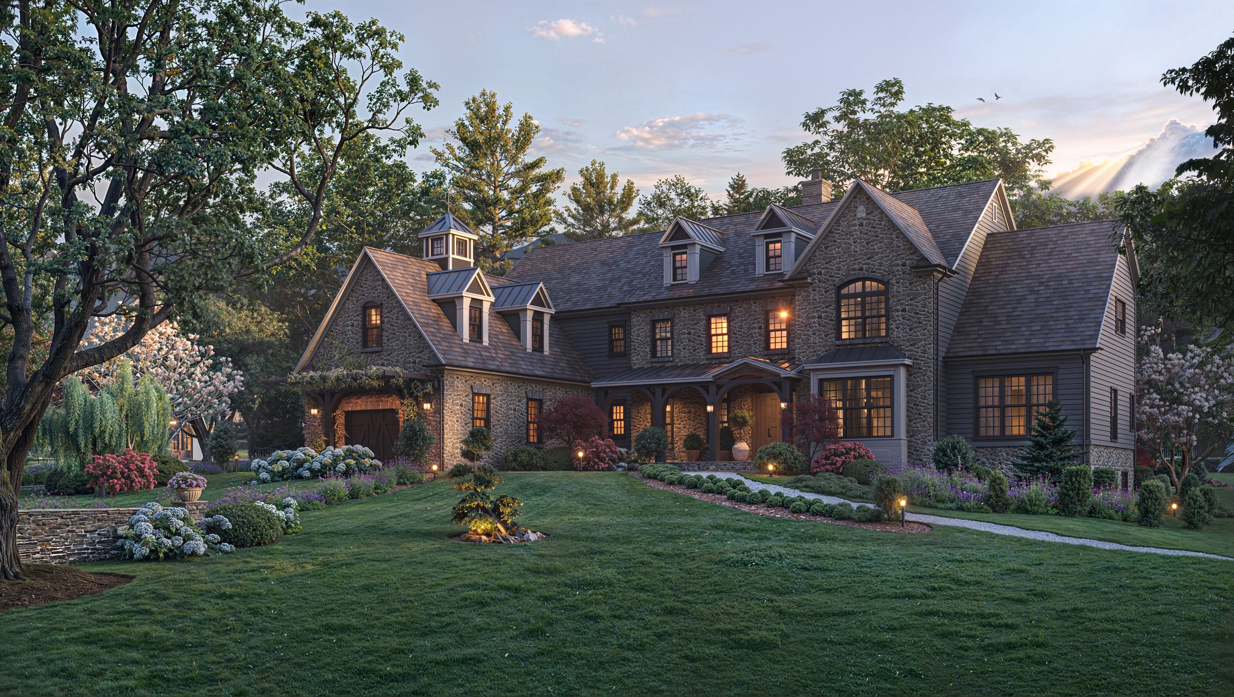 A large, elegant house with stone and gray siding exterior, surrounded by manicured lawn, colorful flowers, and trees, with outdoor lighting at dusk.