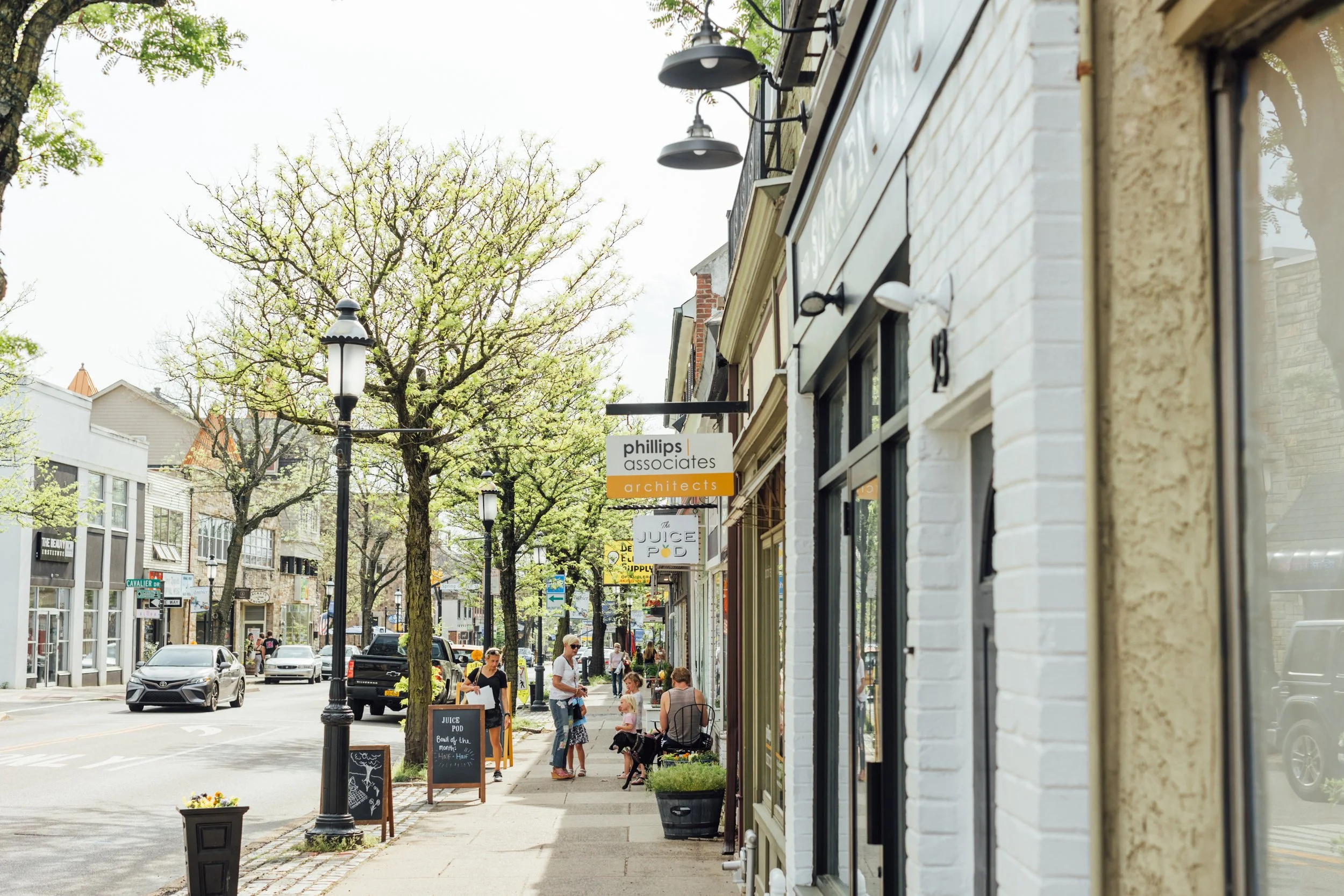 A sidewalk in a small town or city with people walking, sitting, and chatting. Storefronts with signs, trees with budding leaves, and parked cars along the street.
