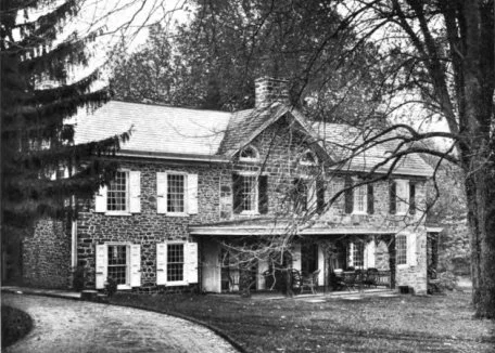 Black and white photo of a large brick residential house with multiple windows, shutters, and a front porch surrounded by trees.