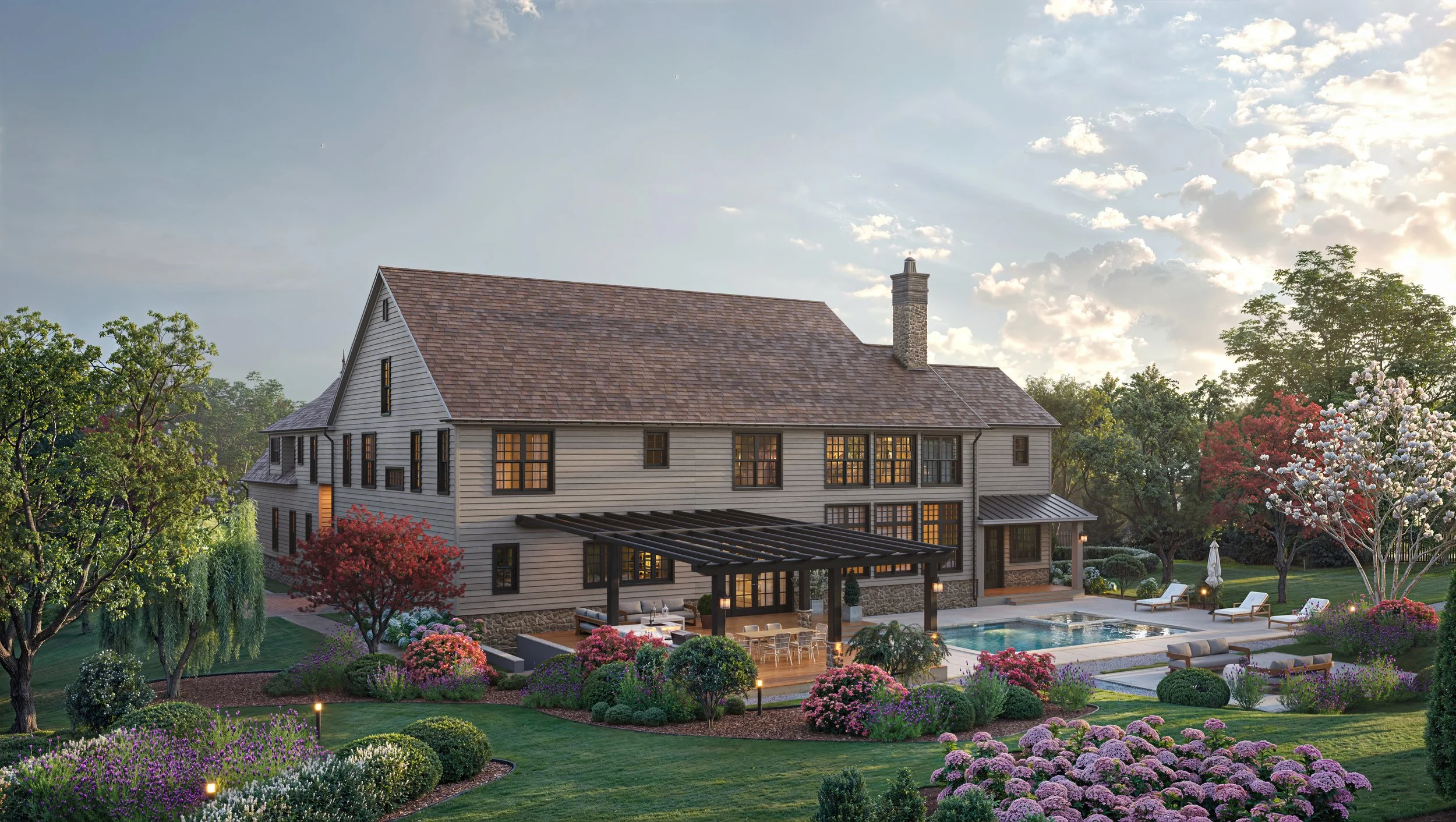 Large house with multiple windows, chimney, and an outdoor patio and pool, surrounded by colorful flowering bushes and trees, under a partly cloudy sky.
