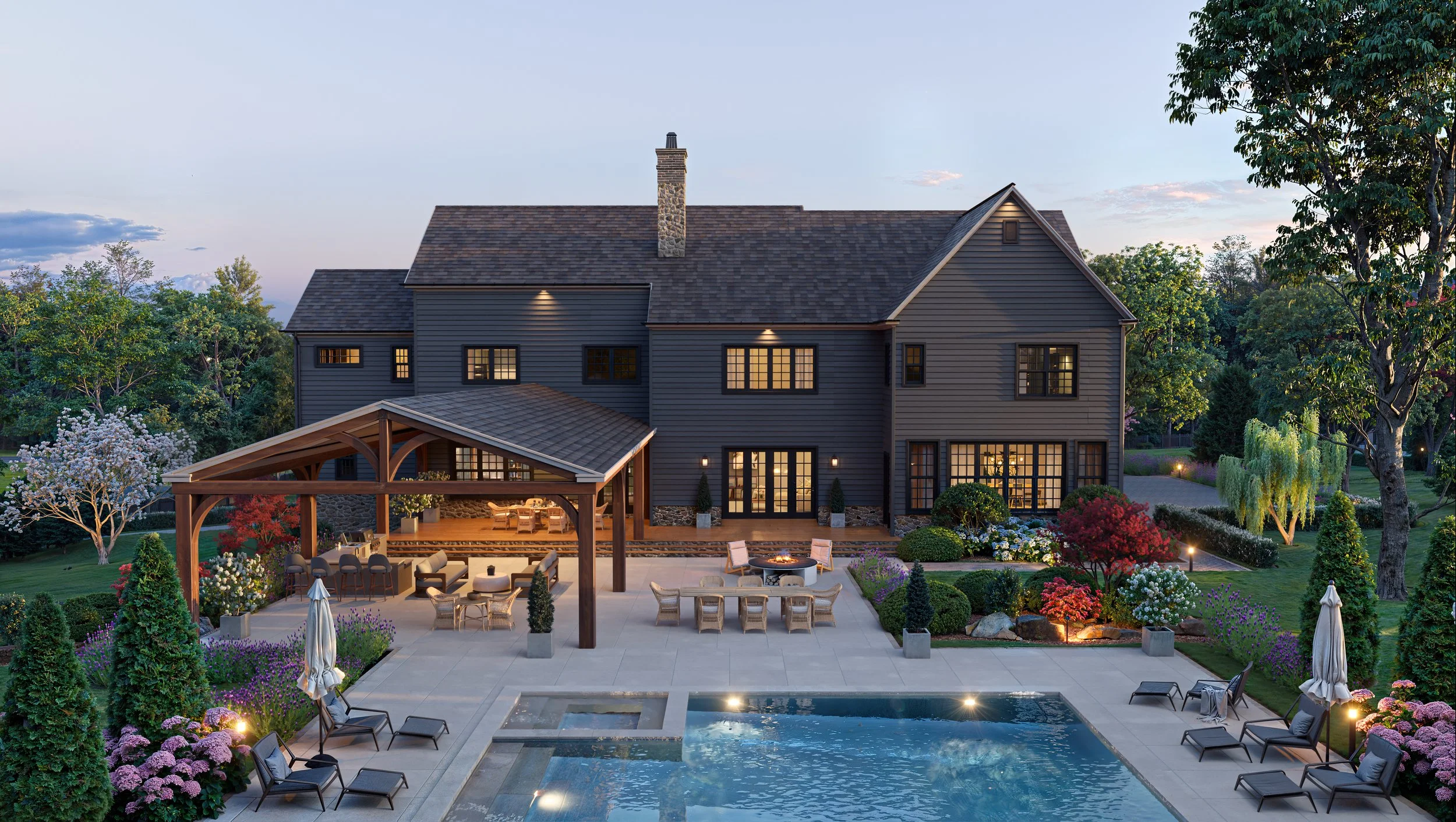 A large, dark blue house with multiple windows, surrounded by lush trees and colorful flowering plants, during evening with outdoor lighting, featuring a spacious patio with seating areas, a pool, and a wooden pergola.