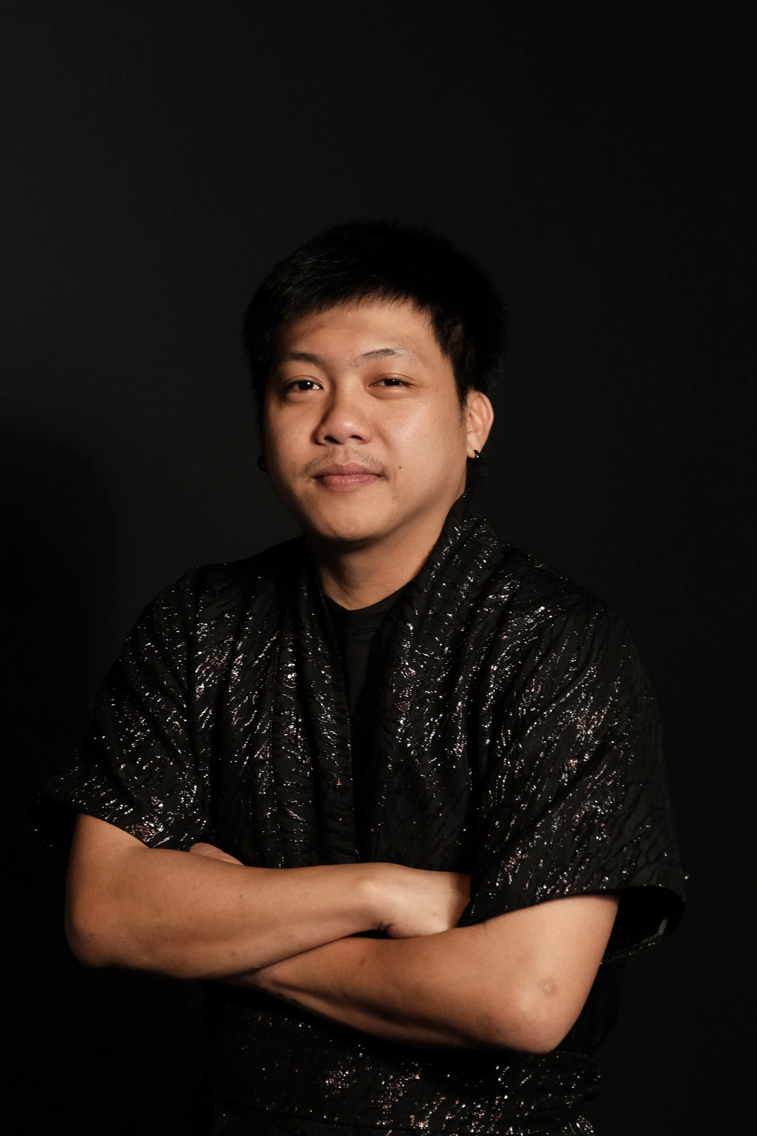 A young man with short black hair, wearing a black, shiny, textured shirt, poses with arms crossed against a black background, looking at the camera with a slight smile.