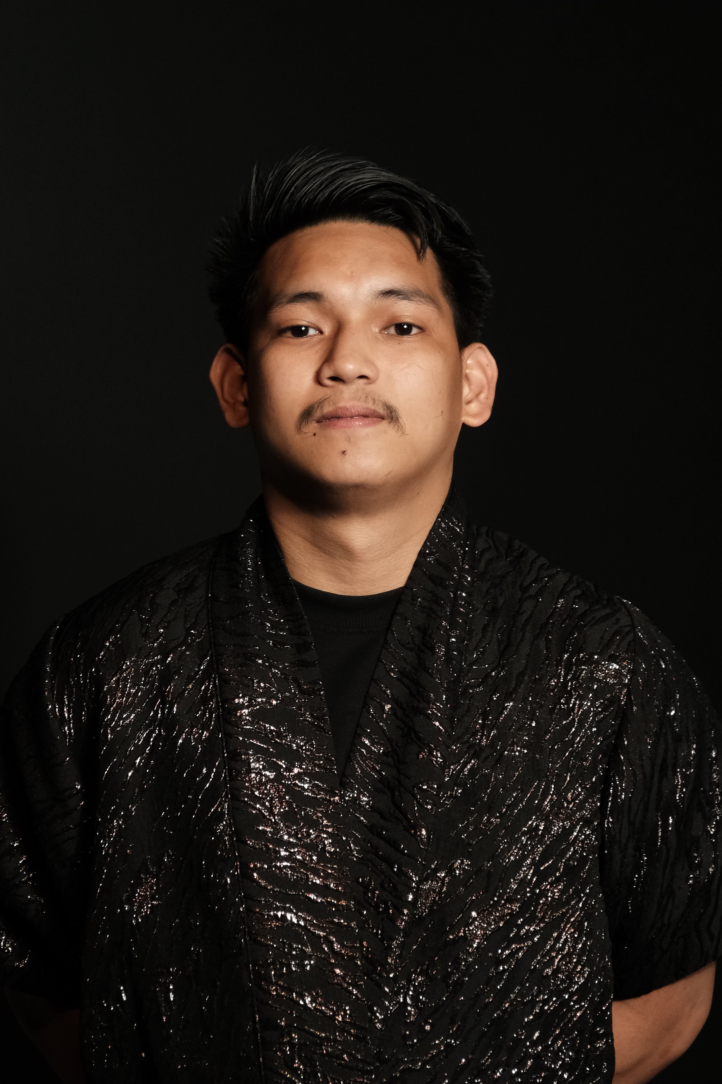 A young man with black hair styled upward and a beige mustache, wearing a black textured jacket with shiny, crinkled patterns, stands against a black background.