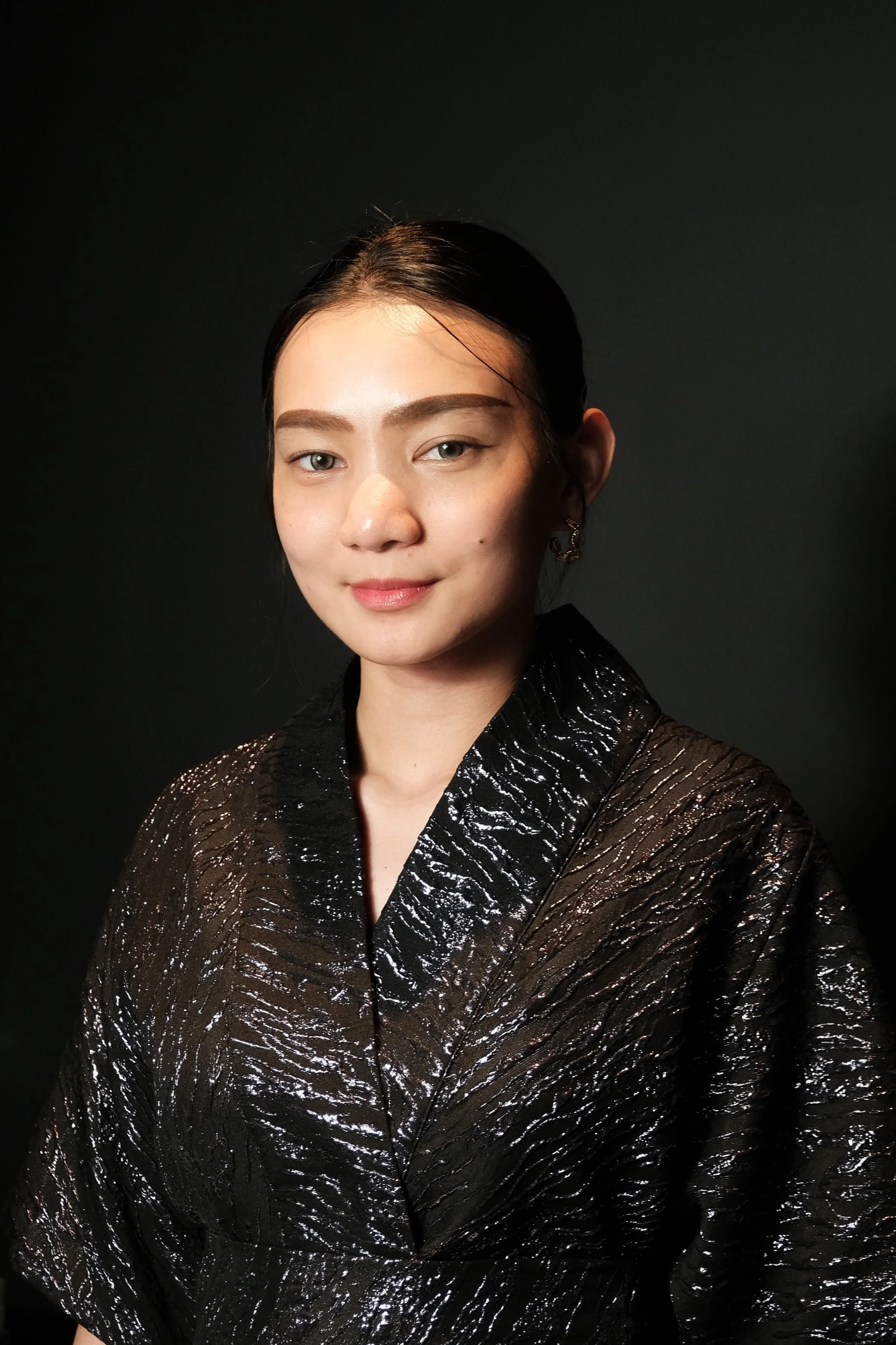 A young woman with dark hair styled back, wearing a black, shiny, textured blouse, posing against a dark background.