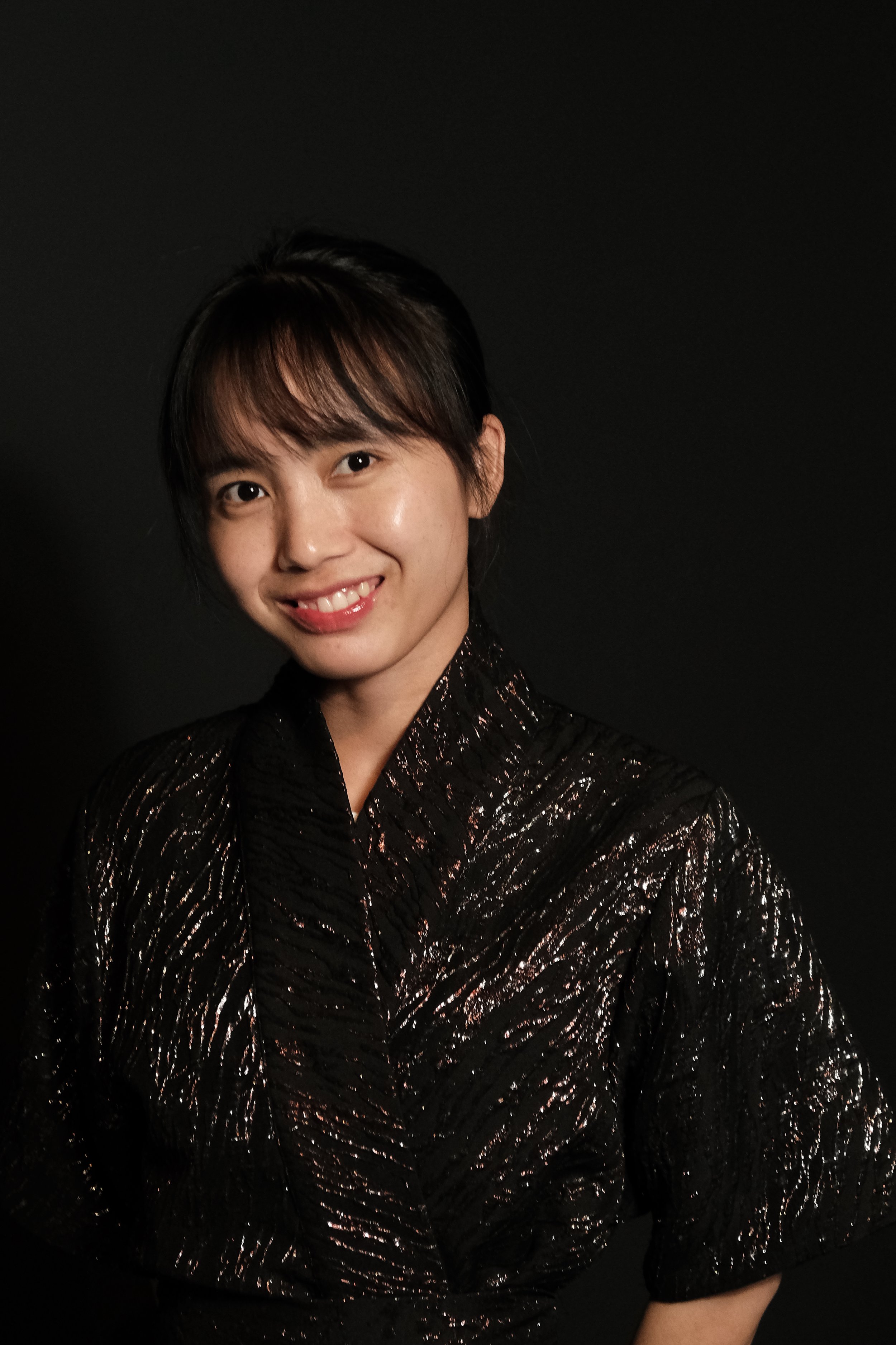 A person with short dark hair smiling against a black background, wearing a shiny, textured black garment.