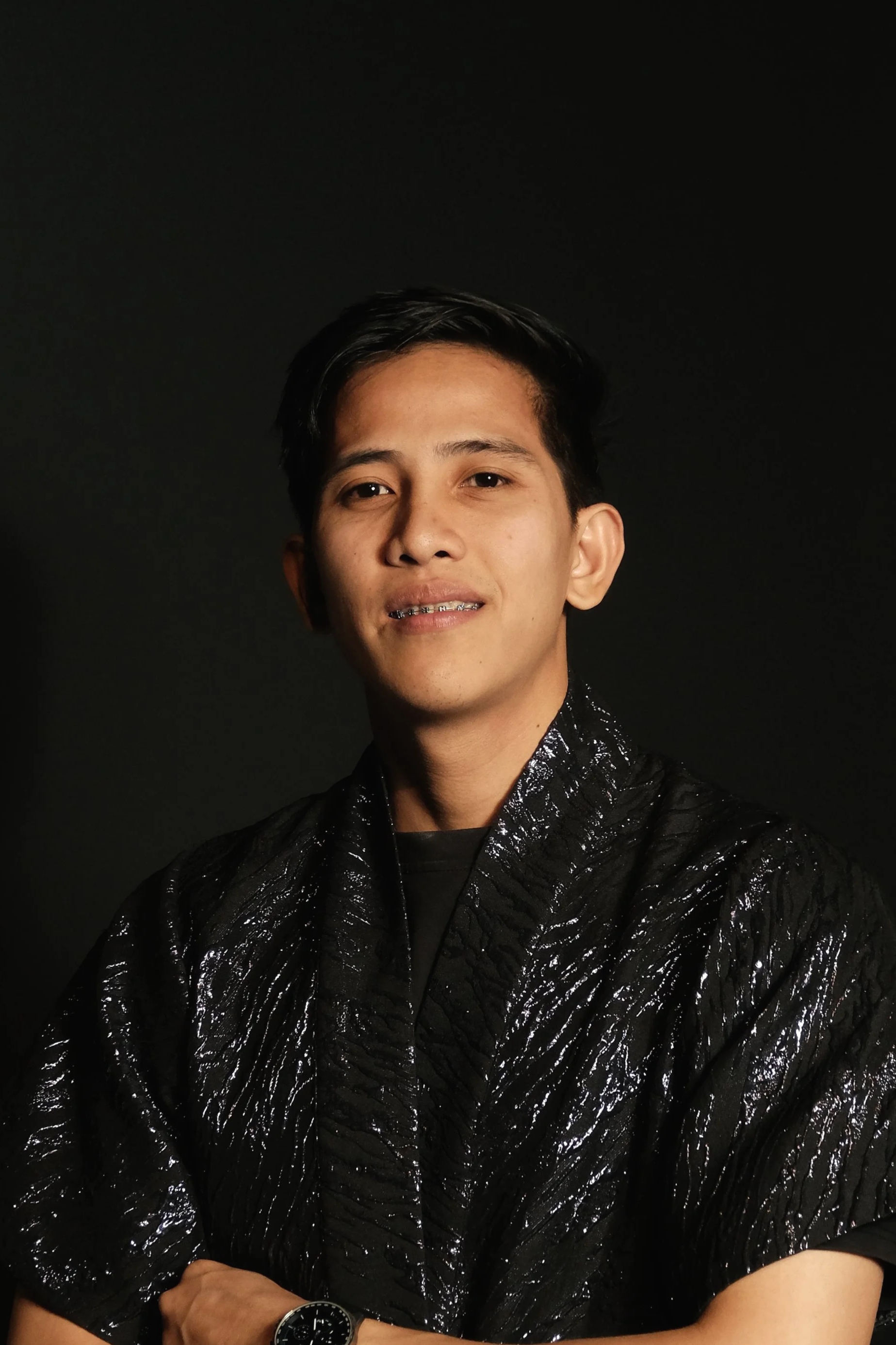 A young man with dark hair wearing a sparkly black jacket, standing against a plain dark background.
