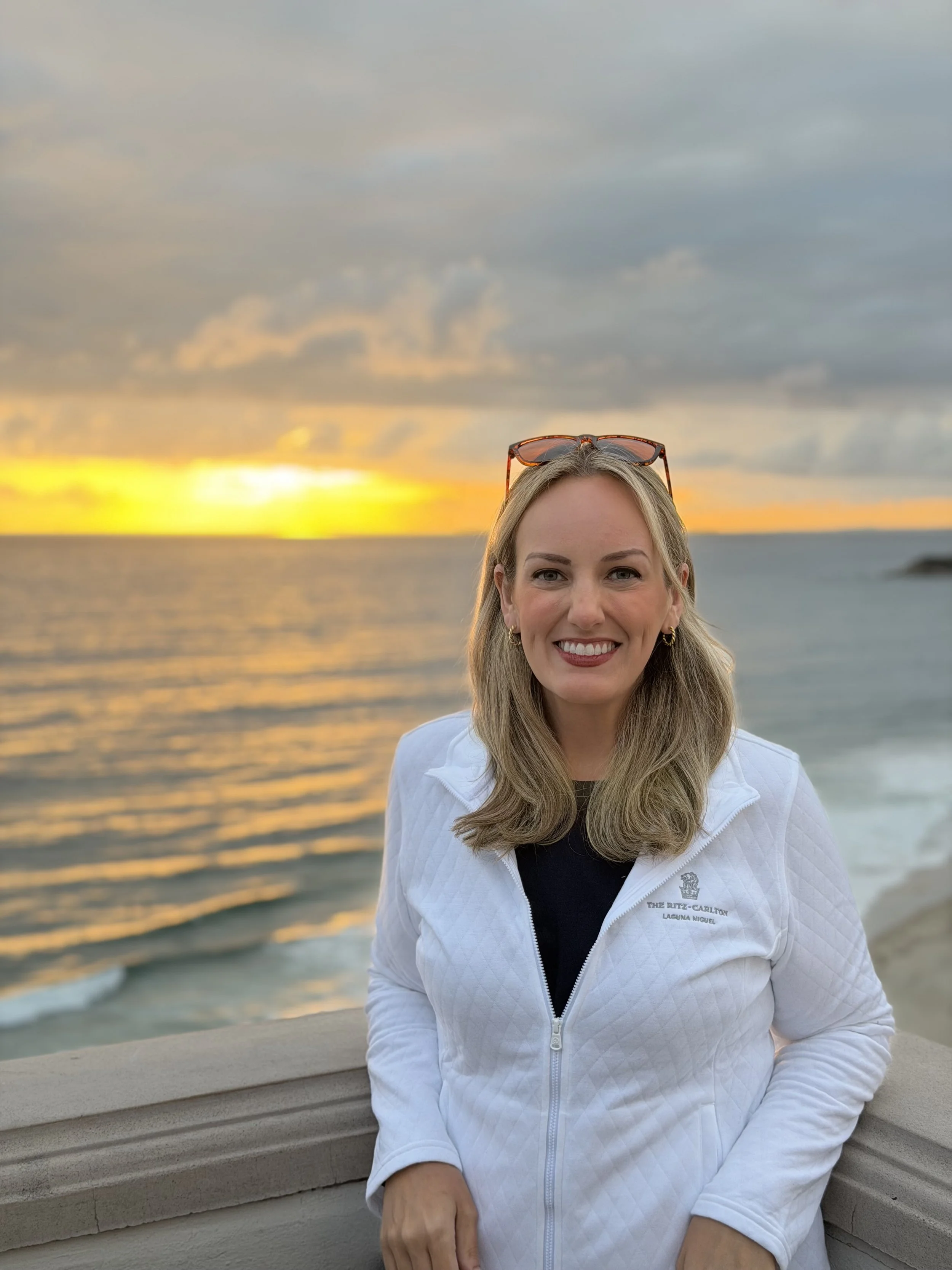 A woman with blonde hair and sunglasses on her head, smiling, wearing a white jacket, standing outdoors near a body of water with a yellow-orange sunset and cloudy sky in the background.