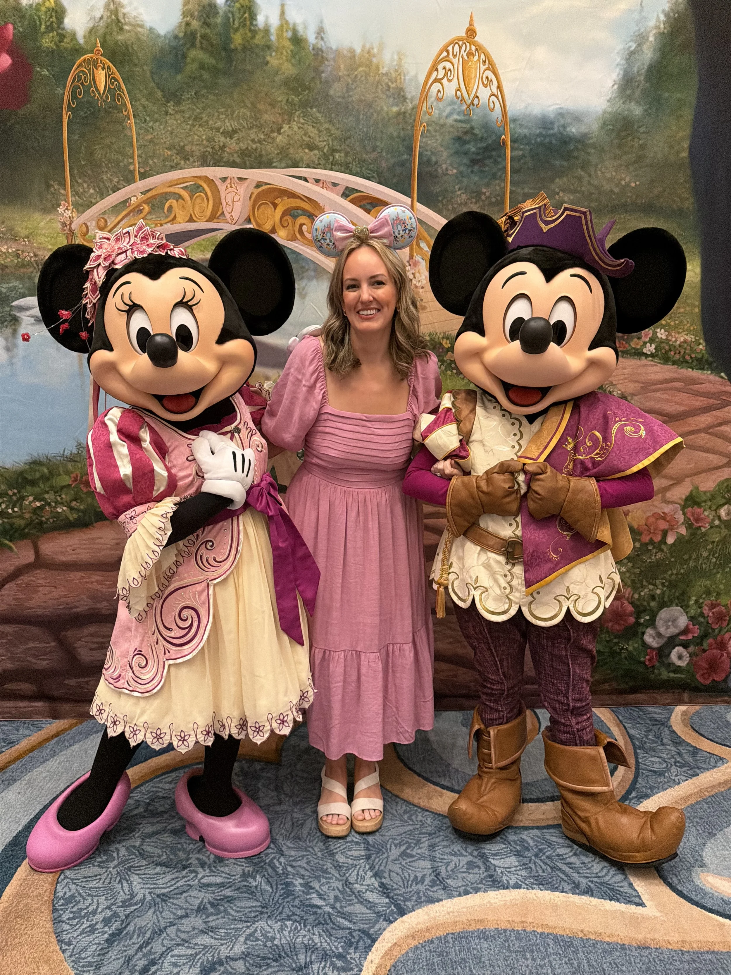 A woman standing between two Disney characters, Minnie Mouse and Mickey Mouse, at what appears to be a themed event or park. Minnie is dressed in a pink and cream dress with a pink bow on her head, Mickey is dressed in a prince outfit with a purple hat. The background is a painted landscape scene, and the floor has a patterned rug.