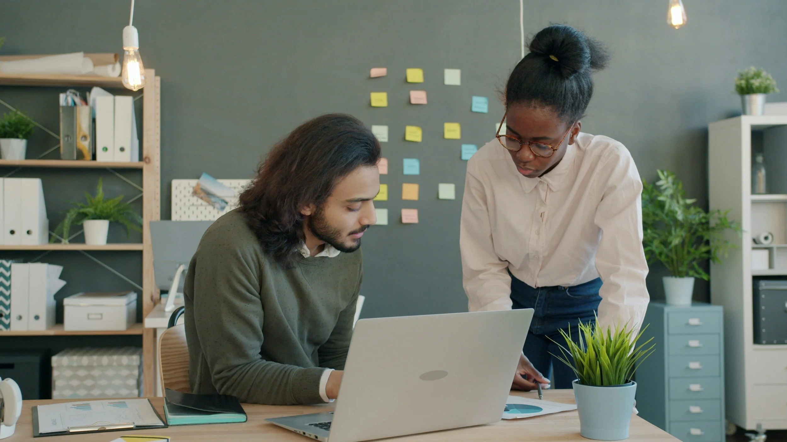 Two coworkers reviewing documents together in a modern office, with a laptop and a plant on the table.
