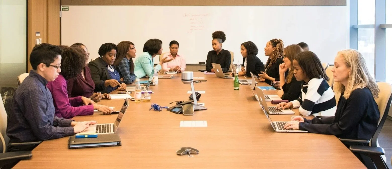 A group of professionals sitting around a long conference table, engaged in a meeting, with laptops and drinks on the table.