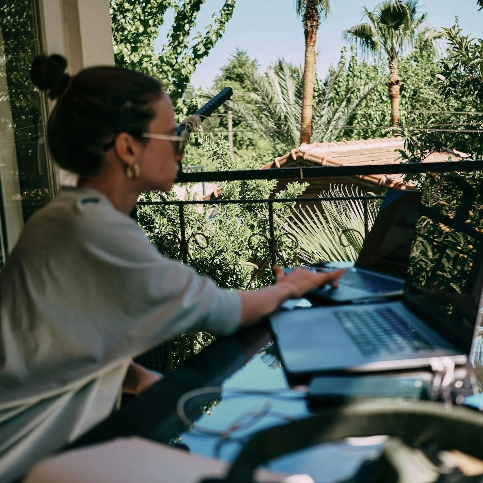 Woman sitting on balcony using a laptop with a lush green garden in the background.