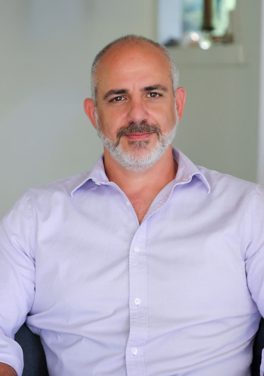 A middle-aged man with a beard and short hair, wearing a light purple dress shirt, sitting and looking at the camera in a room with a blurred light-colored wall and window in the background.