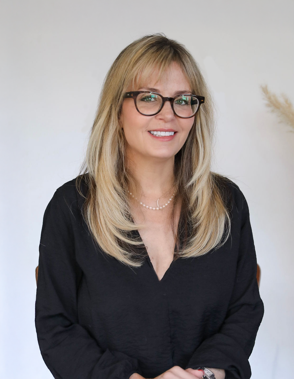 A woman with shoulder-length blonde hair, glasses, and a black top, smiling against a plain white background.