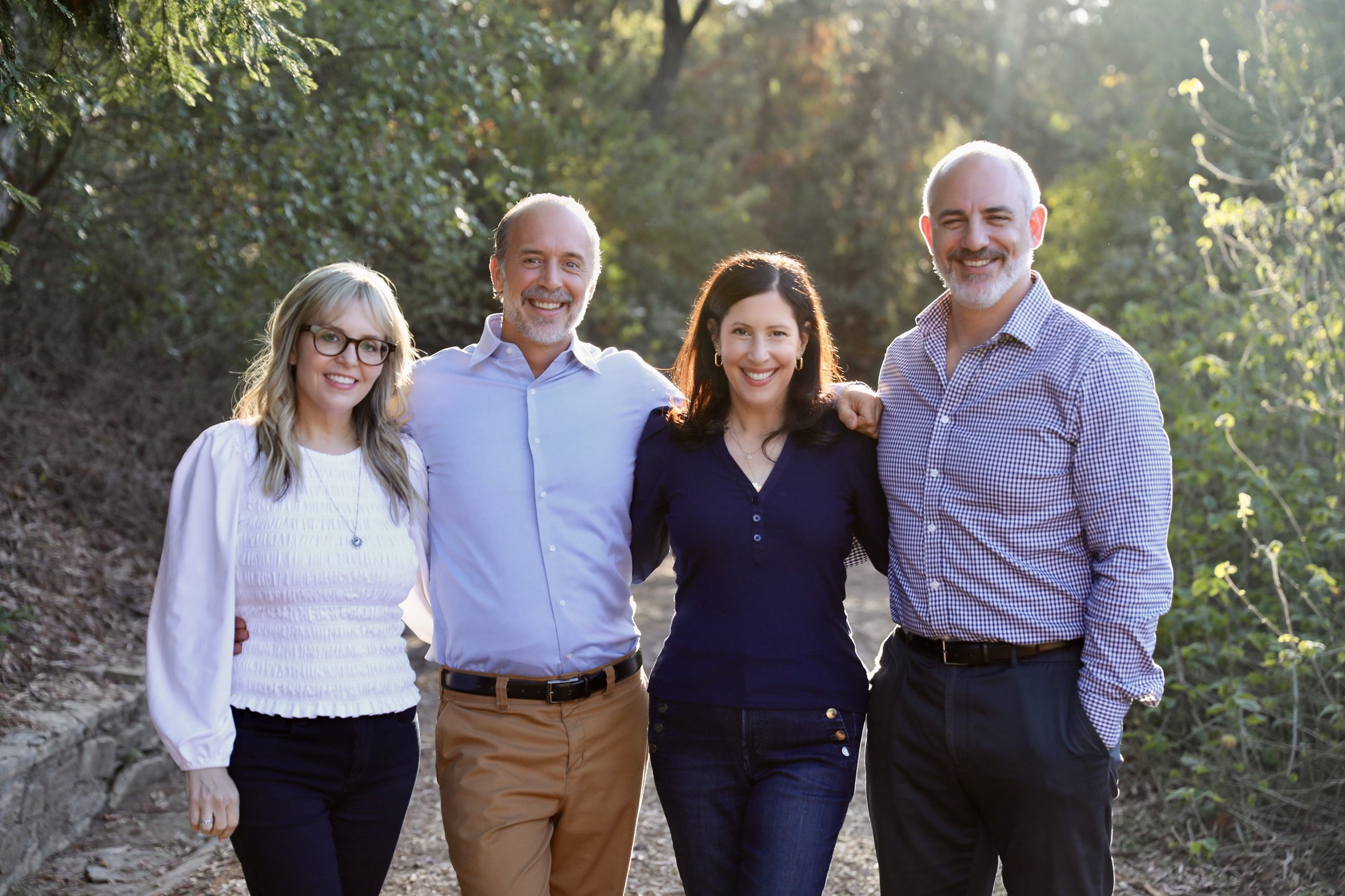 Group of five smiling adults walking together outdoors on a sunny day with green trees in the background.