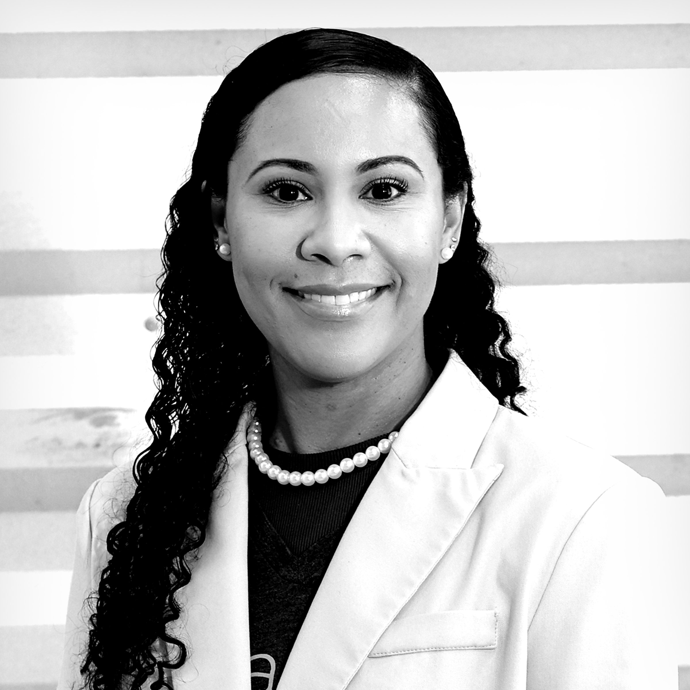 Black and white photo of a woman with long curly hair, dressed in a white blazer, wearing pearl earrings and a pearl necklace, smiling against a striped background.