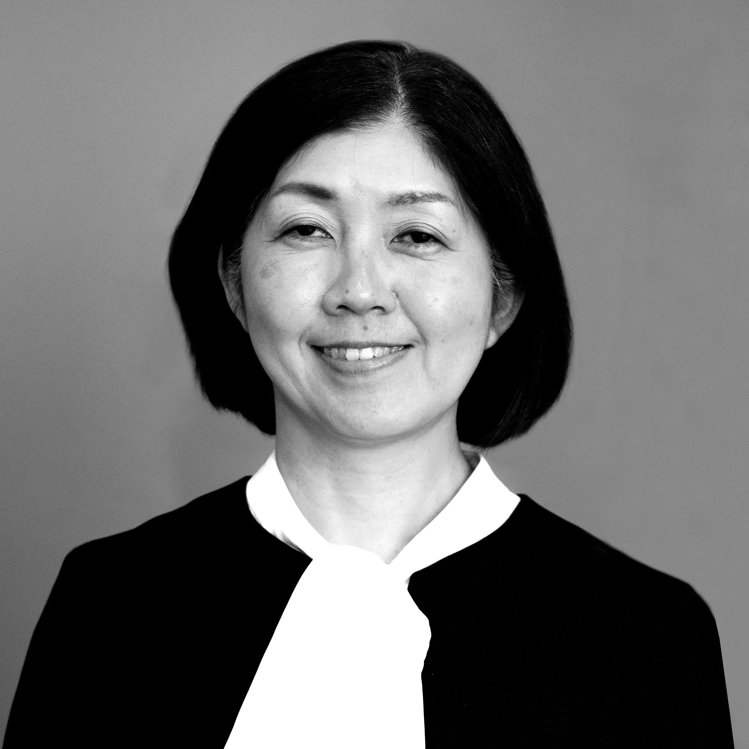Black and white portrait of an Asian woman smiling, wearing a formal outfit with a white collar, against a plain background.