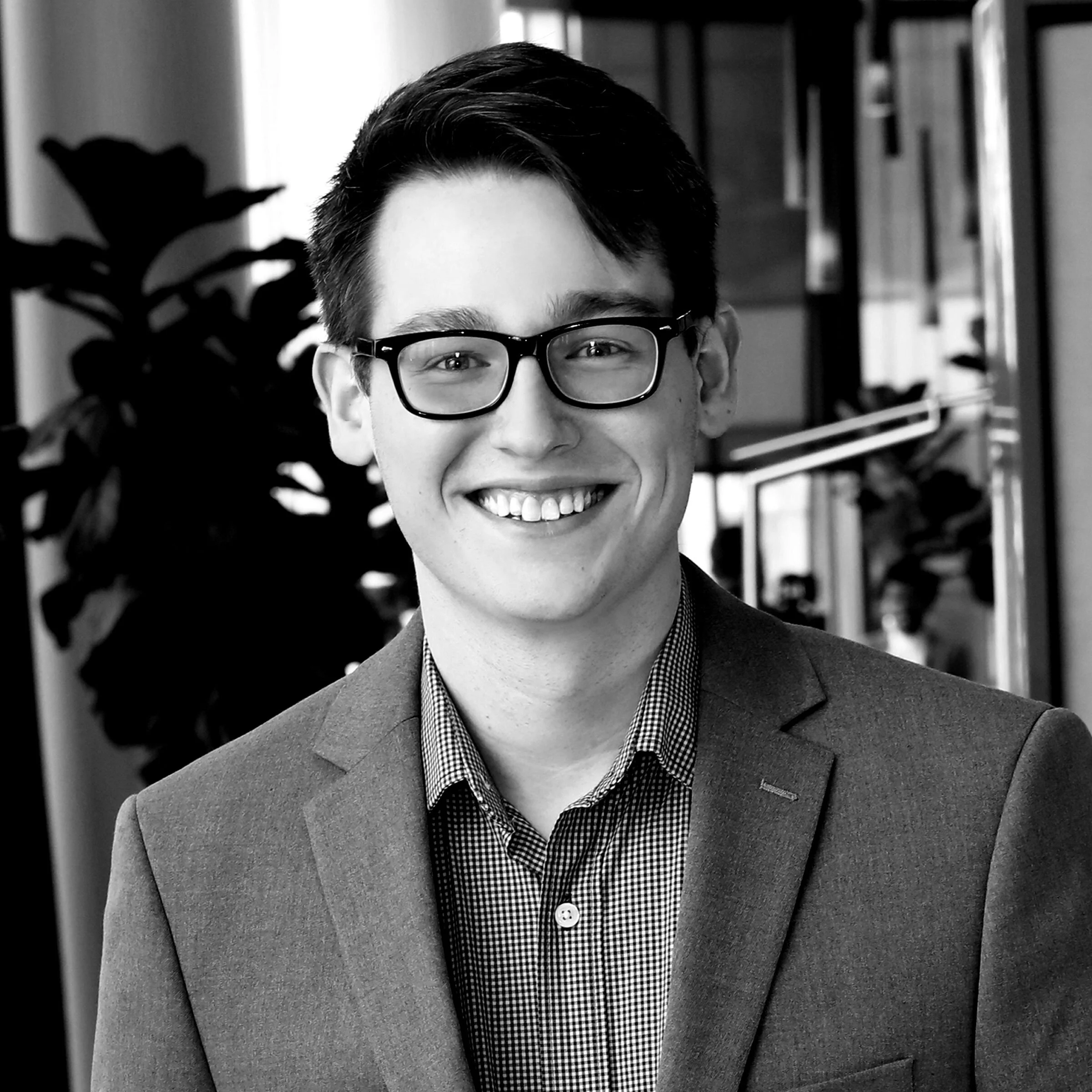 Close-up of a young man with short dark hair, wearing glasses, smiling, in an indoor setting with bookshelves and plants in the background.