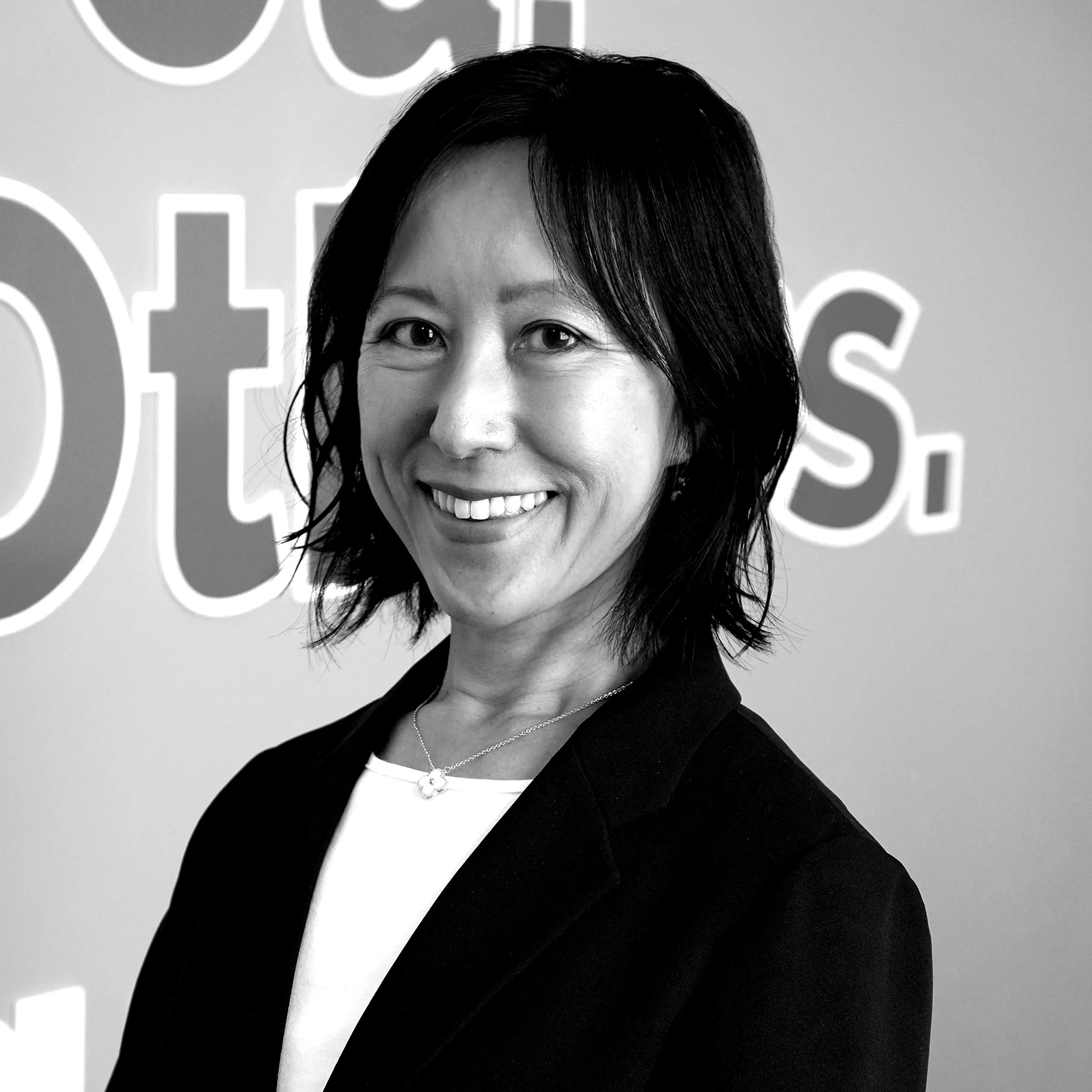 A black-and-white portrait of a smiling woman with shoulder-length dark hair, wearing a necklace with a flower-shaped pendant, and a dark blazer over a white shirt, standing against a background with a large partial logo or sign.