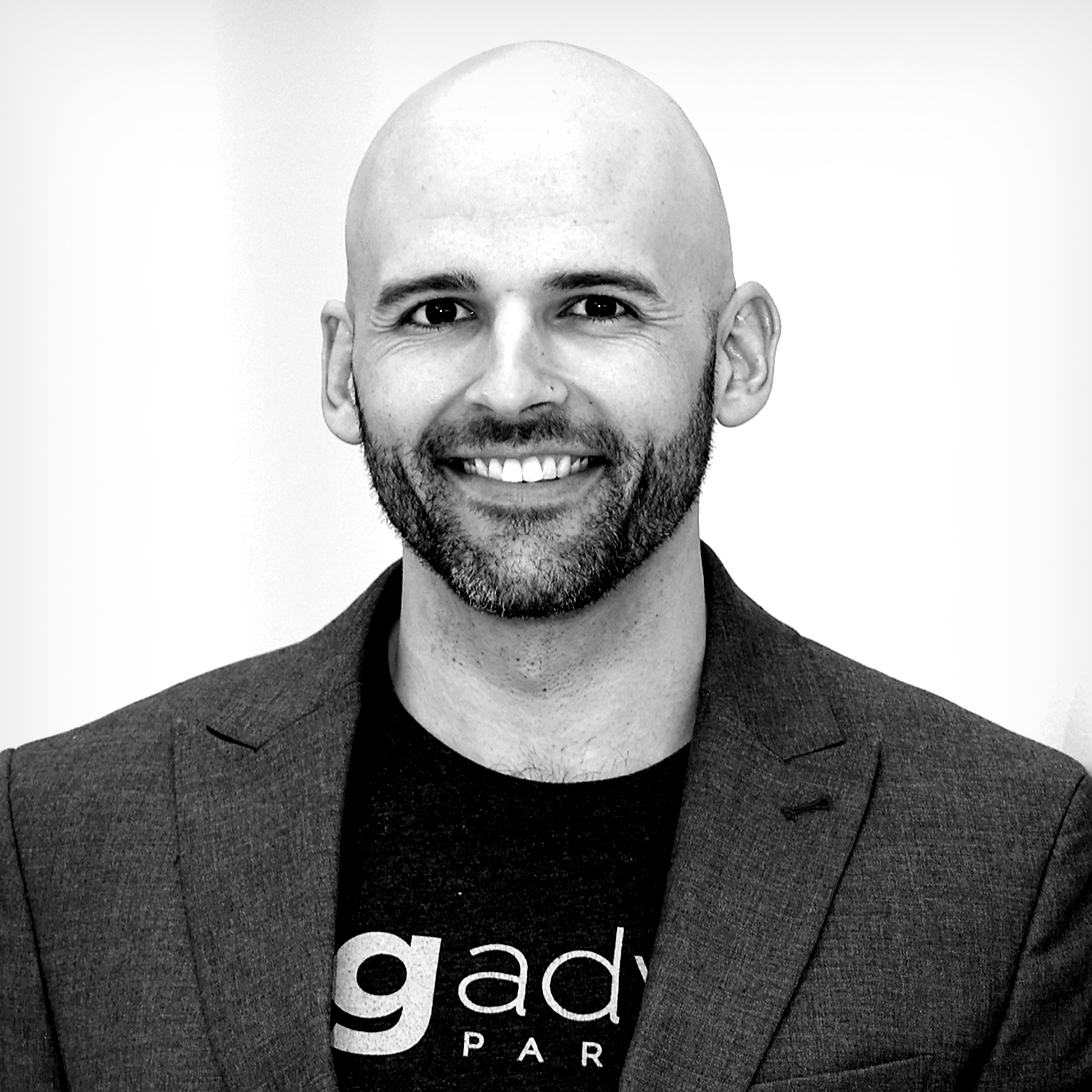 A smiling man with a beard and shaved head wearing a dark blazer and a black t-shirt that says 'dad' with a white 'd' and part of the letters 'g' and 'a' visible, against a plain white background.