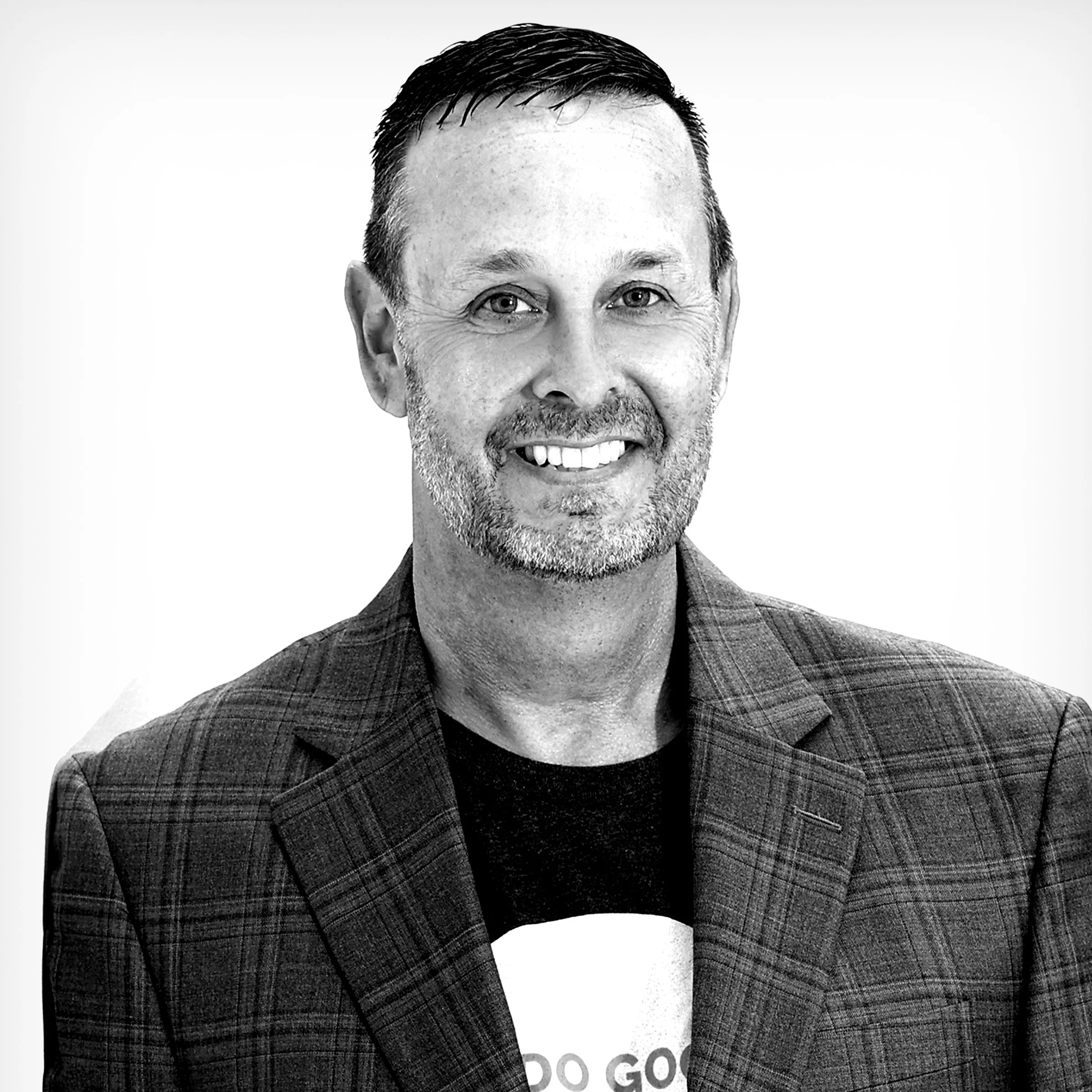 Black and white portrait of a man with short dark hair, a beard, and a friendly expression, wearing a checked blazer and a black shirt, standing in front of a plain background.