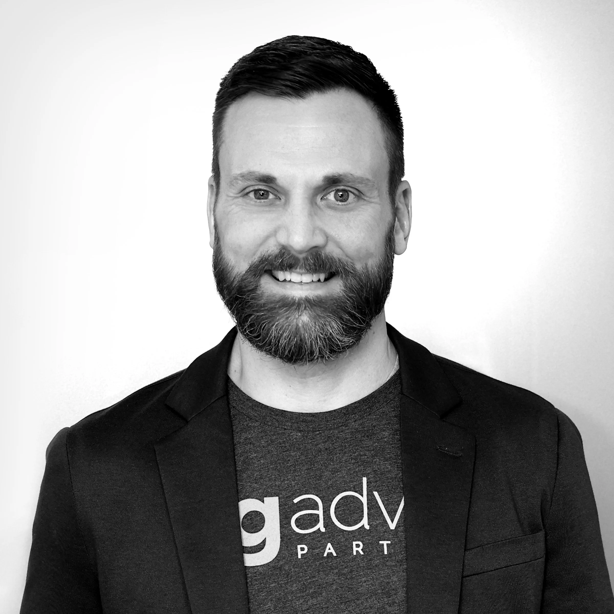 Close-up of a man with dark hair, beard, and smiling, wearing a dark blazer and gray shirt, against a light background.