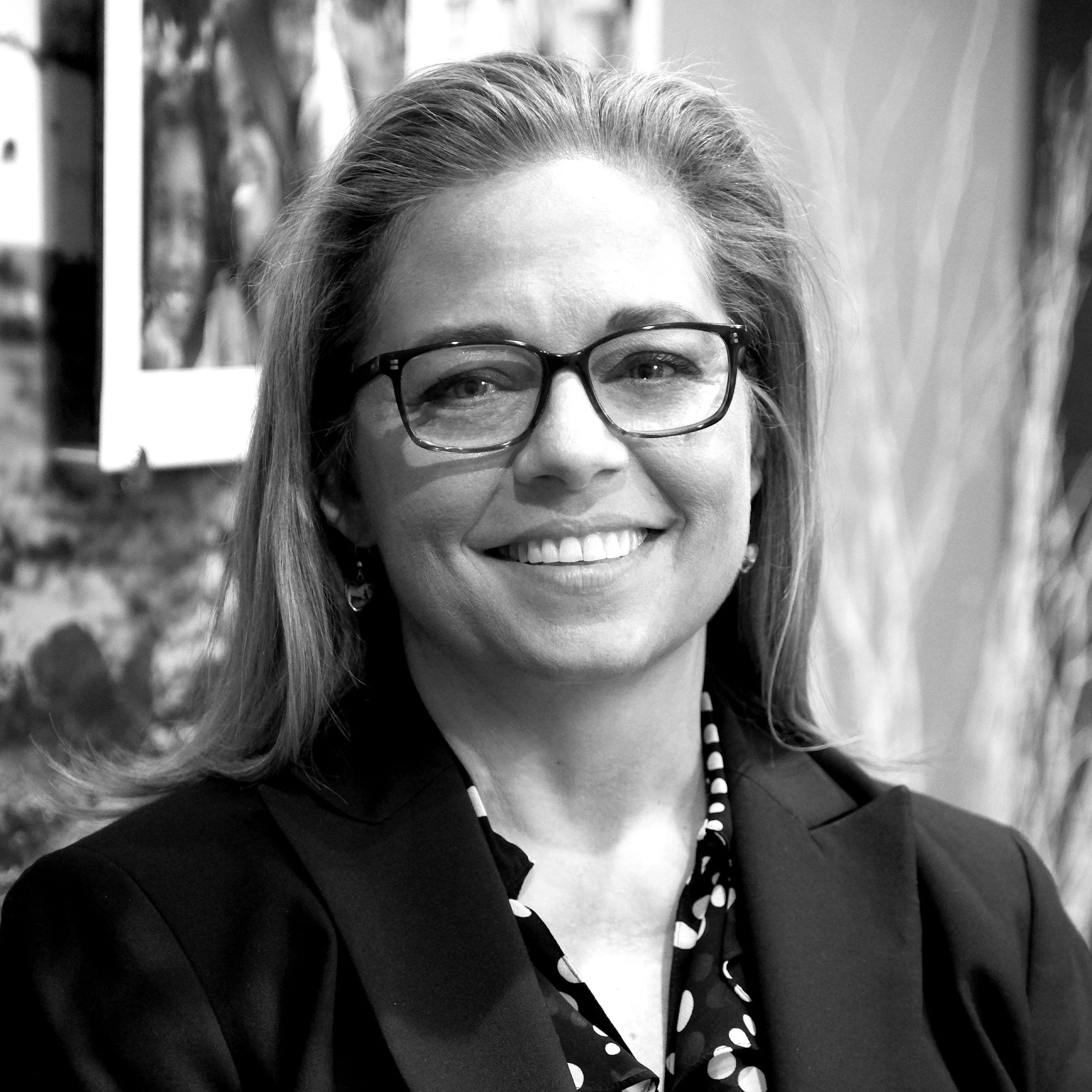 A black and white photo of a woman smiling, wearing glasses, with her hair swept back, and a polka dot shirt.