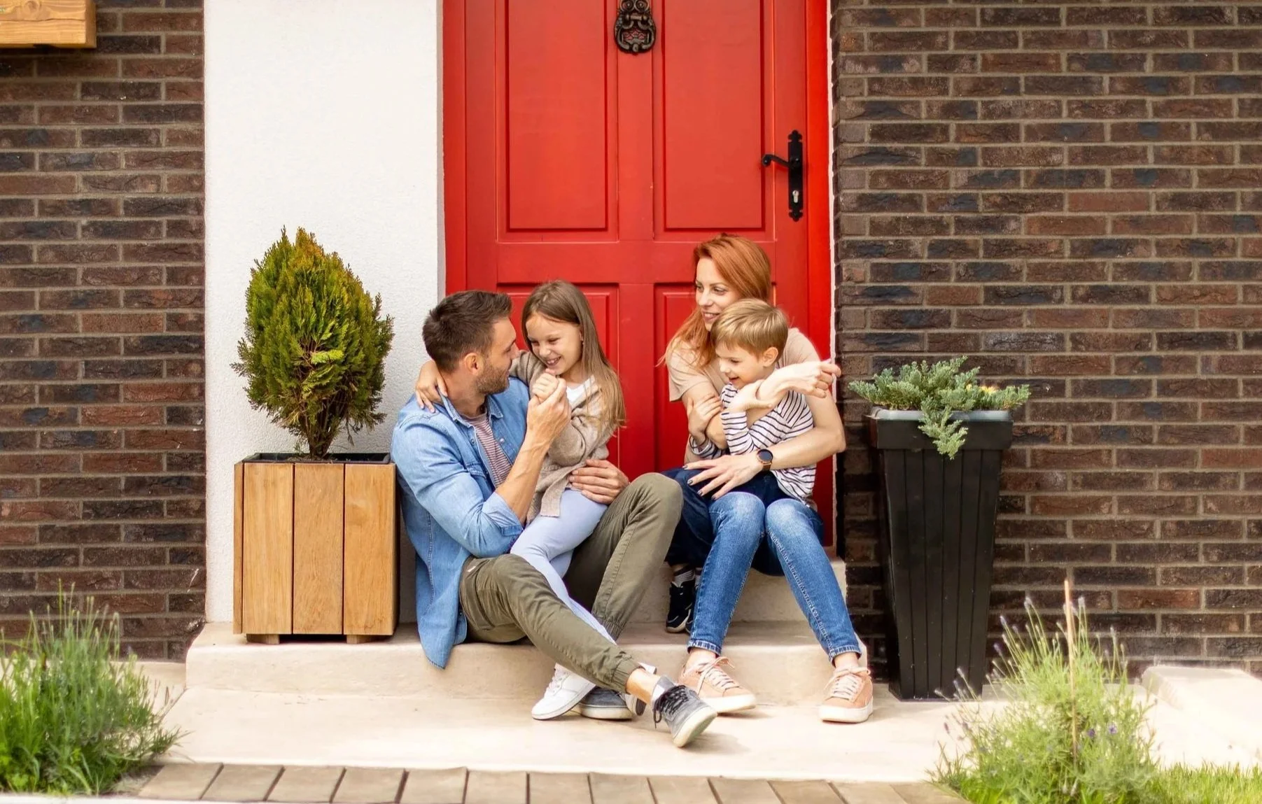 A family of four sitting on the front porch steps of a house. The family includes a man, a woman, and two children. The man and woman are smiling and looking at each other. The children, a girl and a boy, are sitting on the adults' laps. The porch is decorated with two large planters with green plants, and the house has brick walls with a red door.